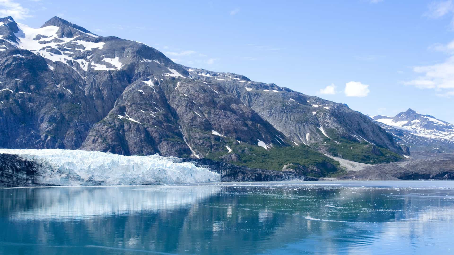 A beautiful landscape of Glacier Bay, Alaska, showing a massive wall of ice and a calm body of water with majestic, snow-capped mountains in the background.