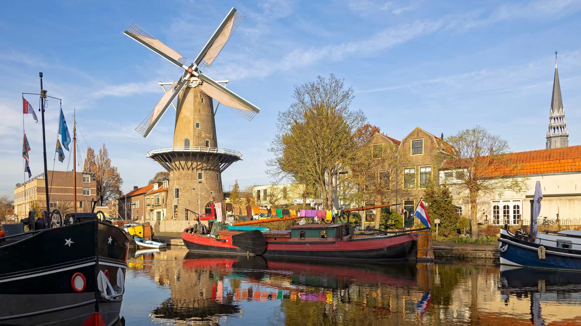 A scenic canal in Gouda, Netherlands, with historic barges moored in the foreground. The 't Slot windmill rises behind a line of houses, and a church spire is visible in the distance.