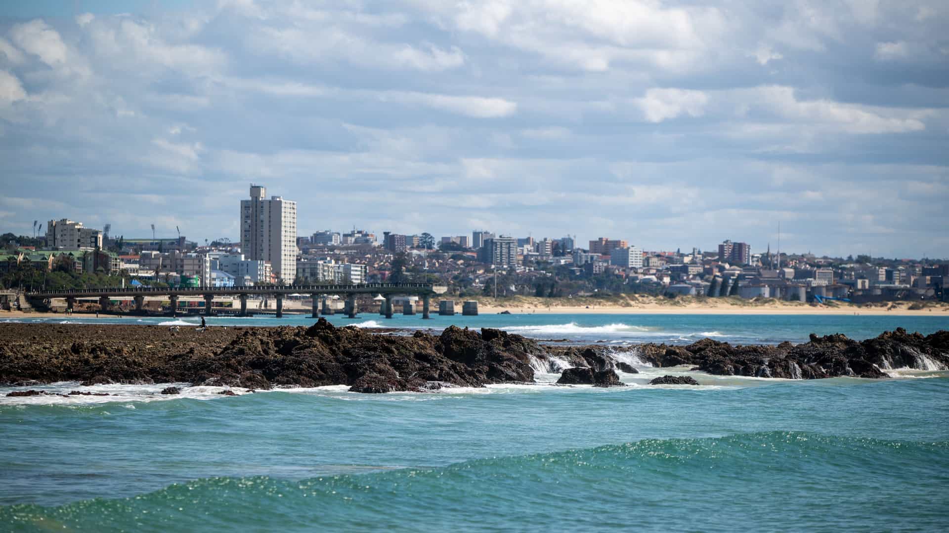 A panoramic shot of the Gqeberha coastline in South Africa, showcasing the city skyline and a pier over the water, with waves crashing on the rocky shoreline in the foreground.