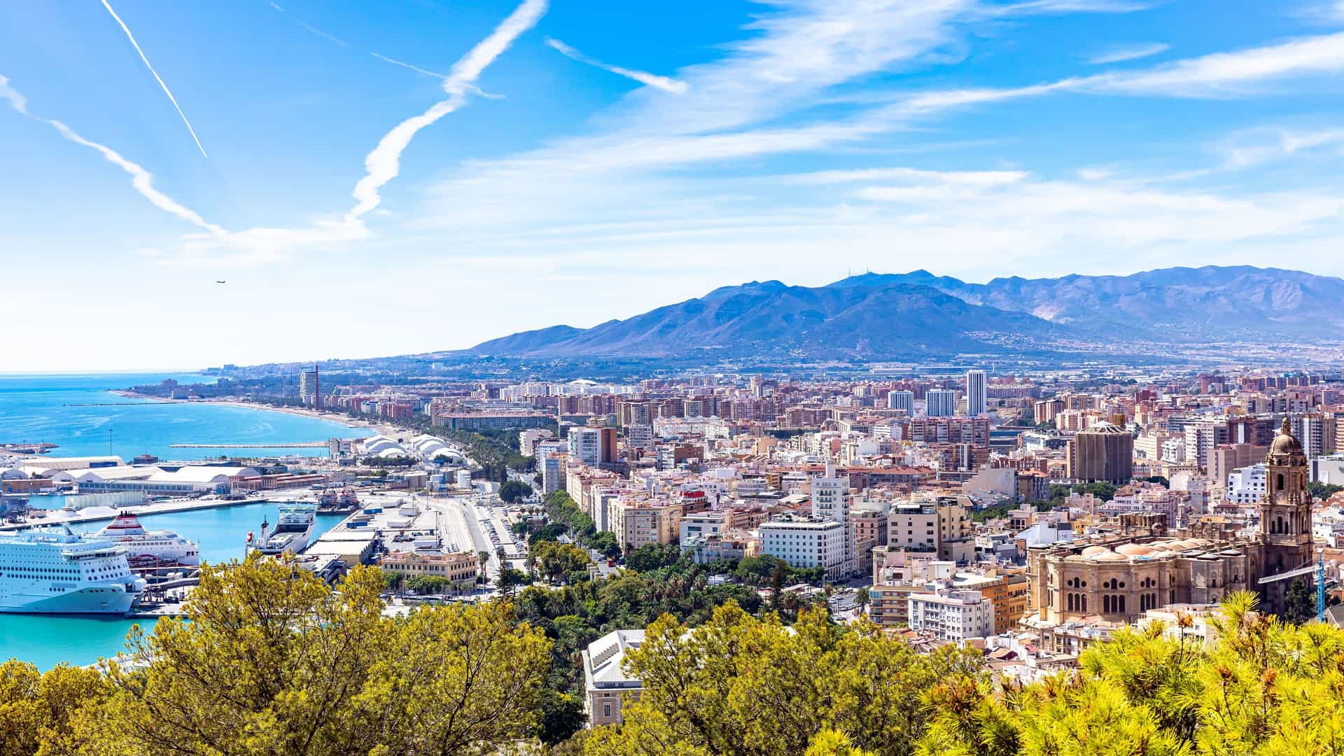 A high-angle panoramic view of Malaga, Spain, with the bustling port and a cruise ship in the foreground, a city skyline, and the beautiful Malaga Cathedral, all set against a backdrop of mountains.