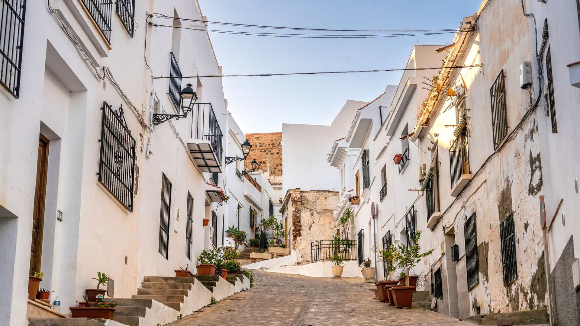 A picturesque view of a narrow, cobblestone street in a historic white-washed village near Motril, Spain, lined with buildings and potted plants.