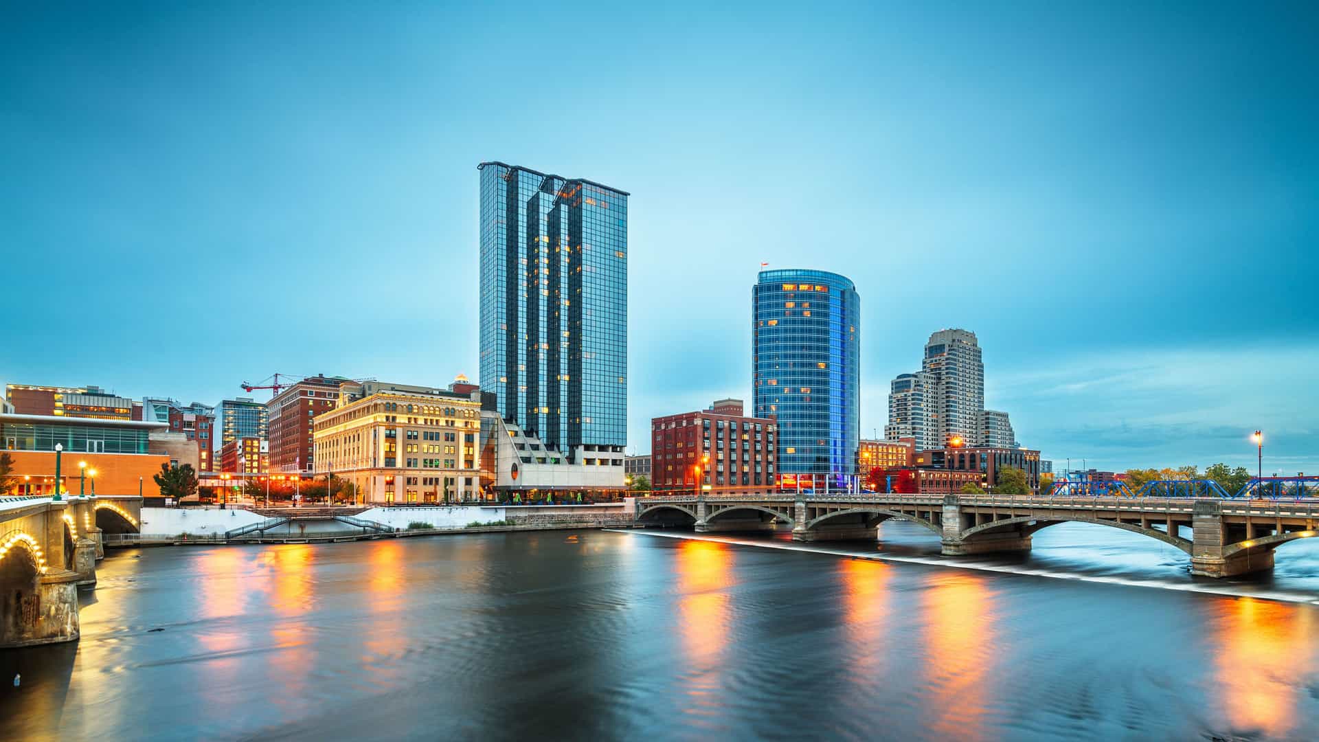 "A panoramic cityscape of Grand Rapids, Michigan, at dusk, with the lights of its downtown buildings and the Gillett Bridge reflecting on the tranquil Grand River.  "