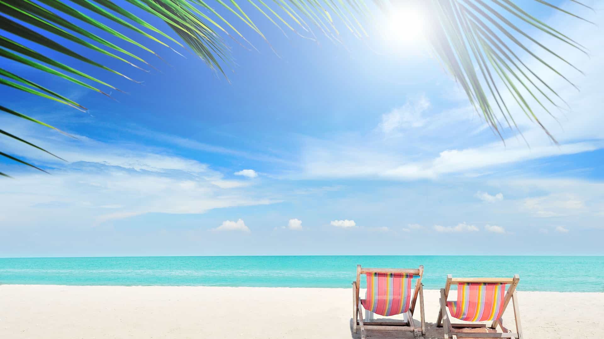 Two colorful striped beach chairs sit on the white sand beach of Great Stirrup Cay, Bahamas, facing the clear turquoise water under a bright blue sky with palm fronds overhead.