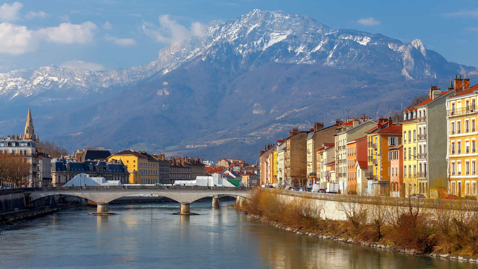 "A scenic view of Grenoble, France, with the Isère River flowing through the city, a bridge connecting the two sides, a row of colorful buildings, and a backdrop of the majestic, snow-dusted French Alps.  "