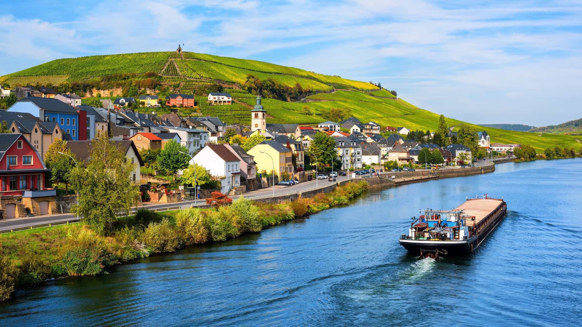 A barge travels down the Moselle River, with the charming town of Grevenmacher, Luxembourg, and the surrounding terraced vineyards on the hillside in the background.