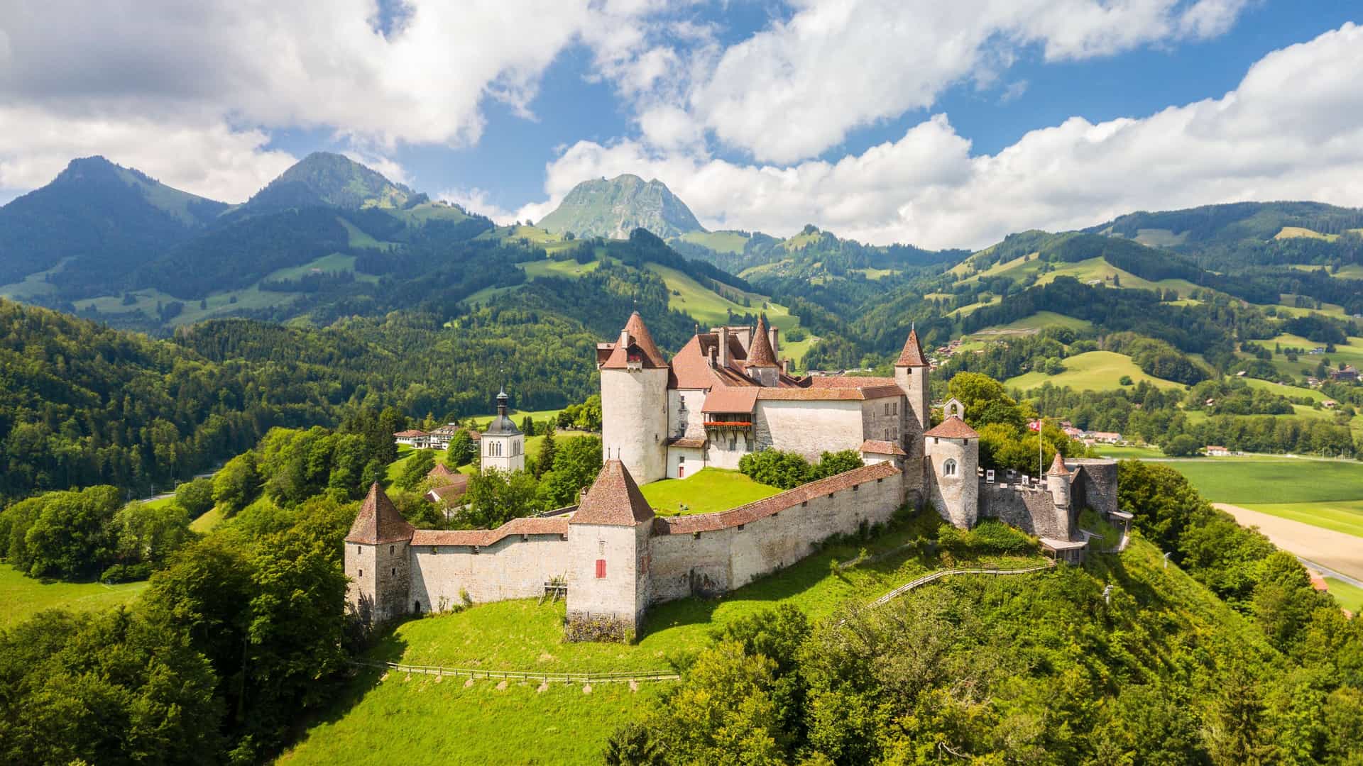 An aerial view of the historic Gruyères Castle, with its red-roofed towers, nestled on a lush green hillside in the scenic Swiss countryside, surrounded by mountains.