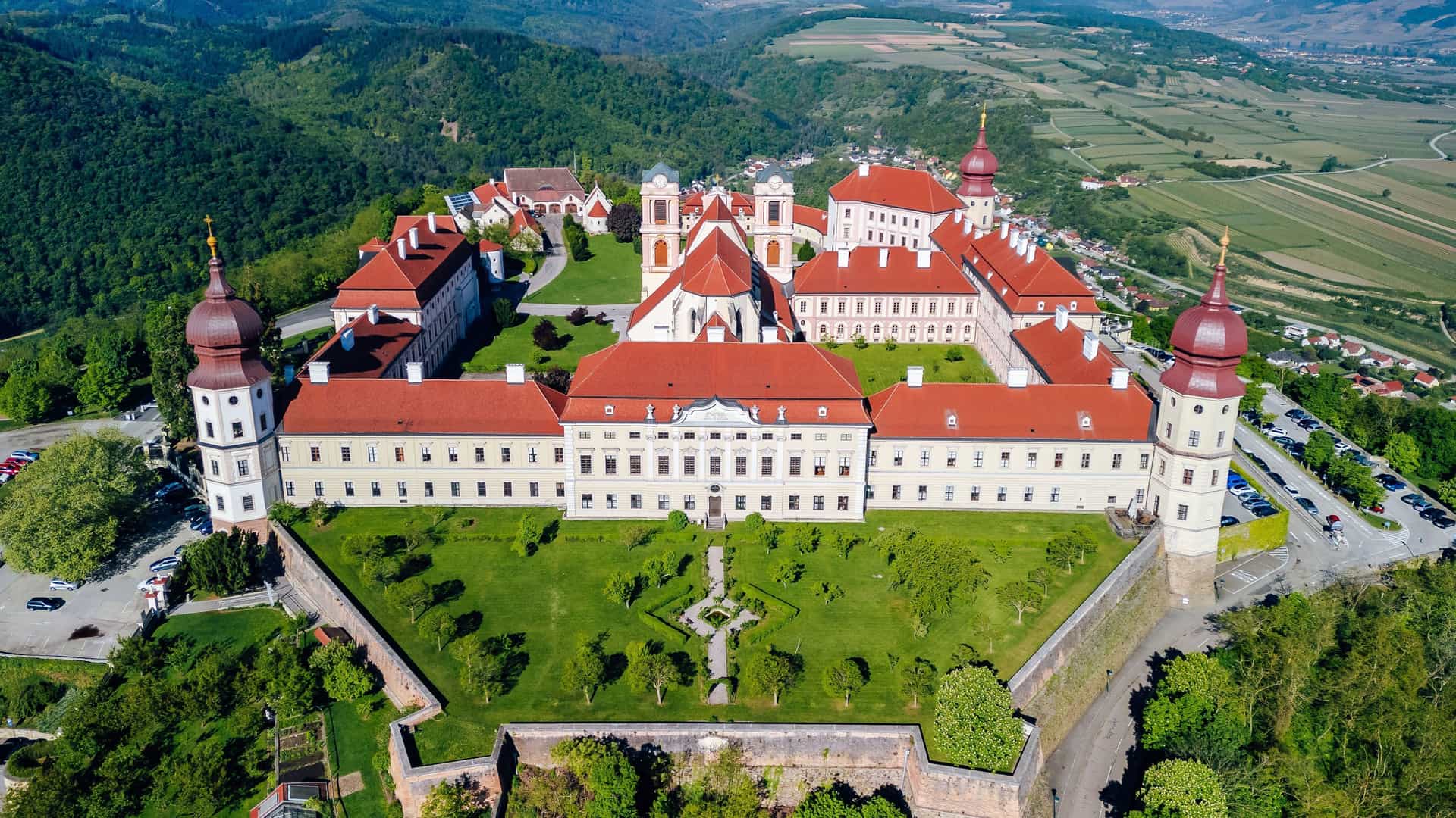 An aerial view of the majestic Göttweig Abbey, a sprawling Baroque monastery with red roofs and onion-domed towers, perched on a hill overlooking Austria's scenic Wachau Valley.
