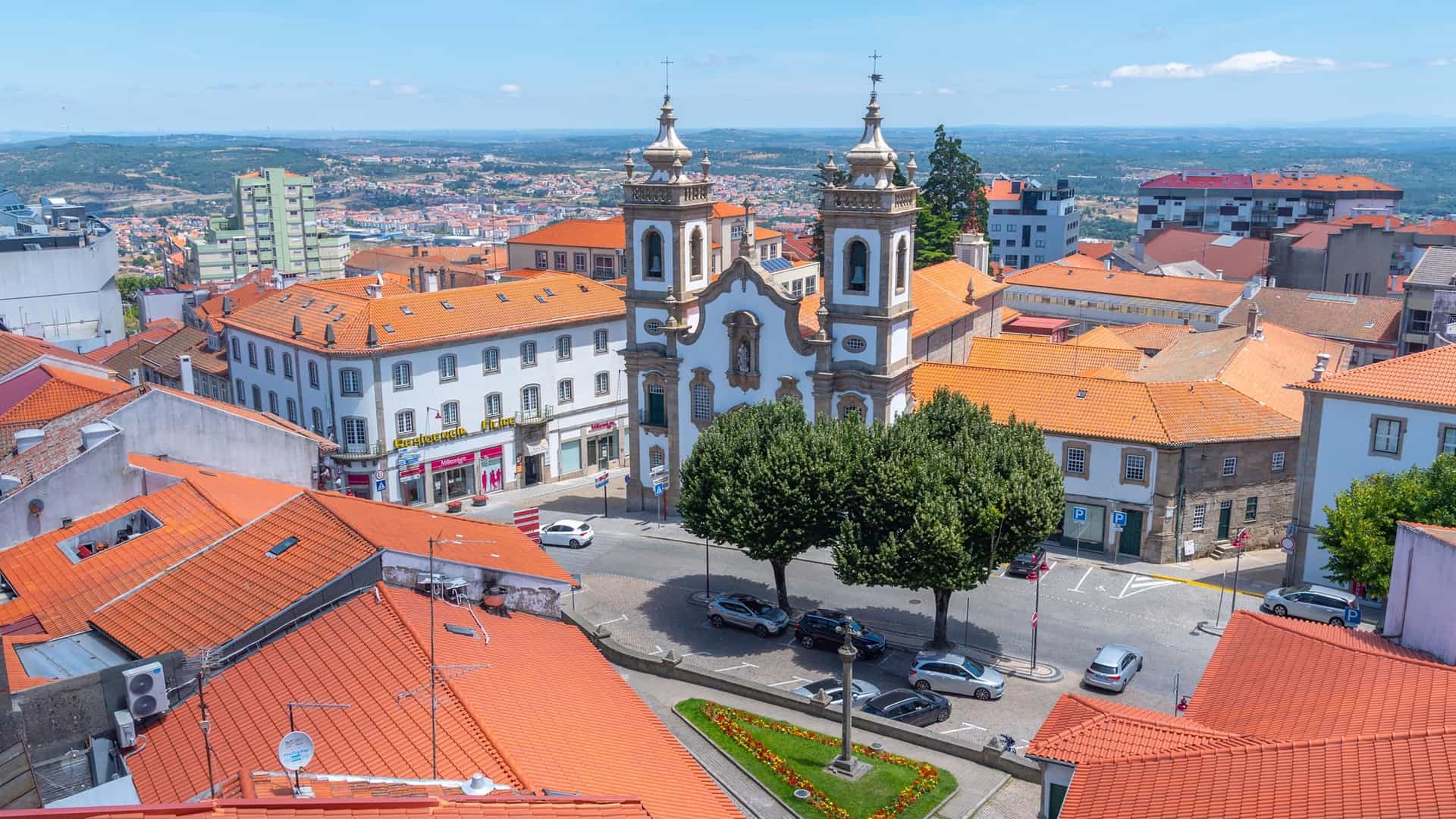 "An aerial view of the historic city of Guarda, Portugal, with a church featuring two ornate bell towers at its center, surrounded by a mix of red-roofed buildings and modern architecture.  "
