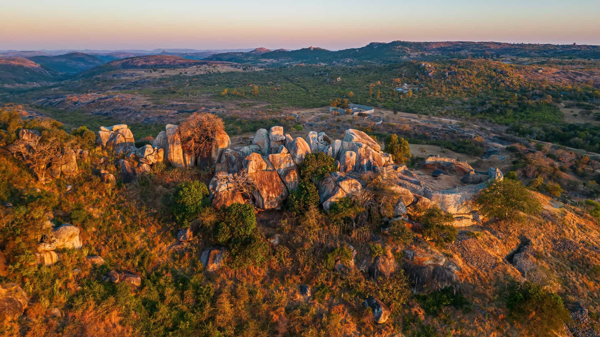 A panoramic view in Gwanda showing ancient stone ruins set among large granite formations and dry vegetation, with warm sunlight casting over the rugged landscape and surrounding hills.