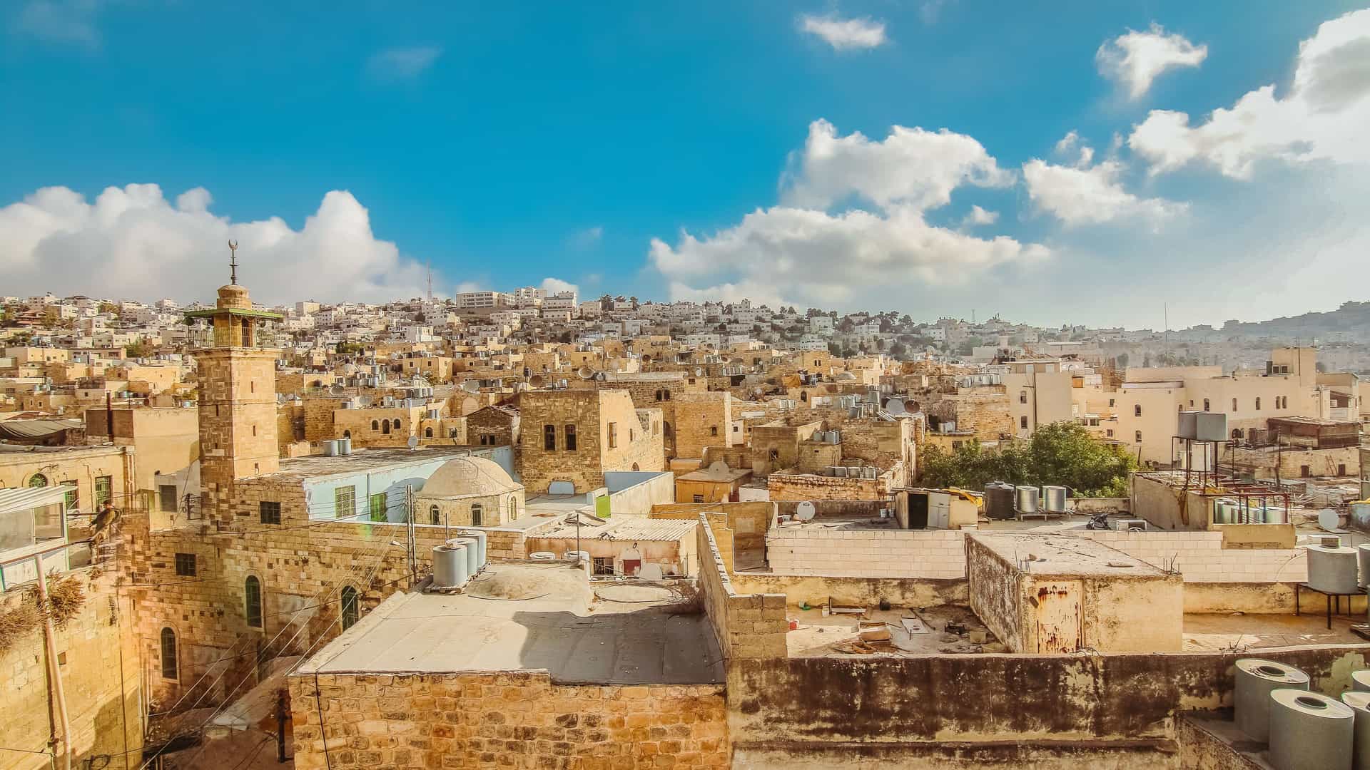 A scenic view of the city of Hebron, Palestine, with the ancient city and its unique architecture sprawling across the hills under a bright blue sky with scattered clouds.