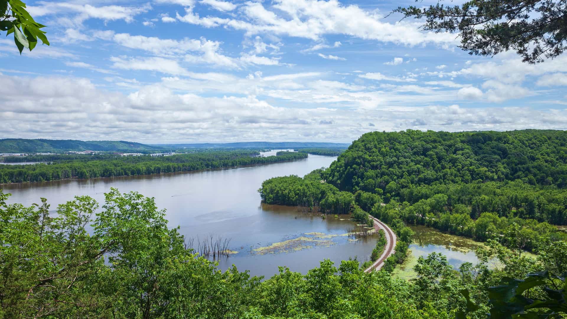A panoramic, high-angle view of a wide river with a forested island in the middle, and a winding road leading to it. The sky is bright blue with scattered clouds.