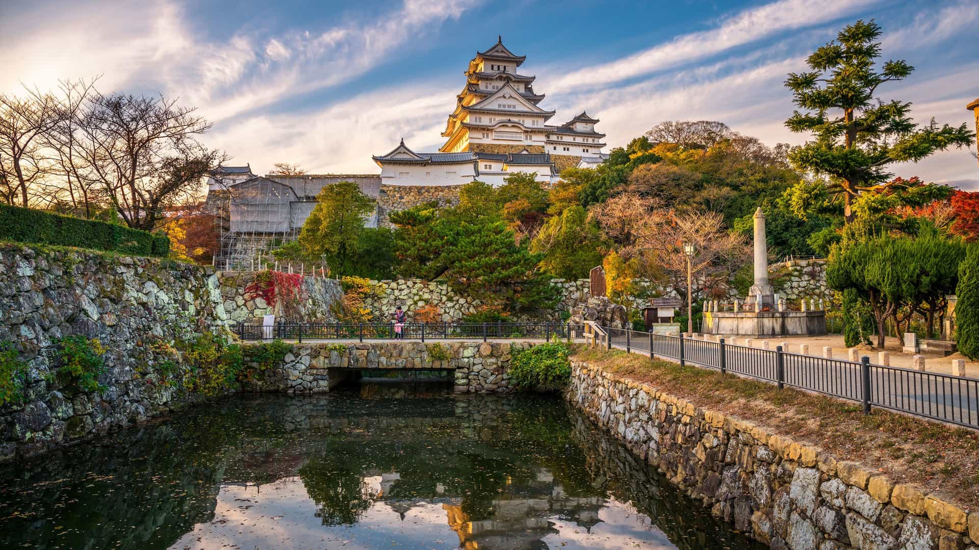 A wide-angle view of the Himeji Castle in Japan, with its majestic white structure towering over a lush green landscape, a moat, and a stone bridge in the foreground.