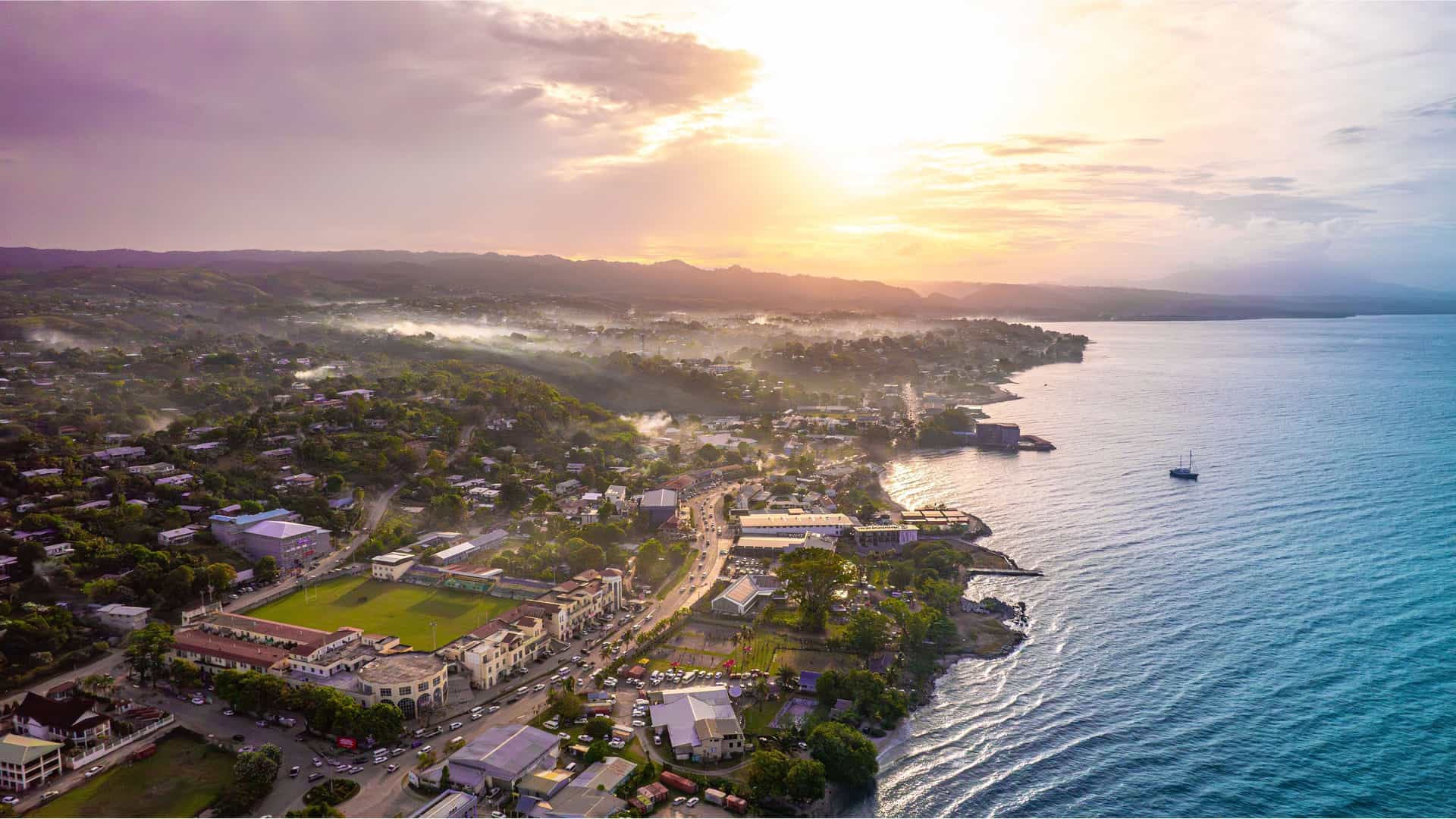 An aerial view of Honiara, the capital of the Solomon Islands, at sunset, showing the city's coastline, buildings, and a lush green landscape.