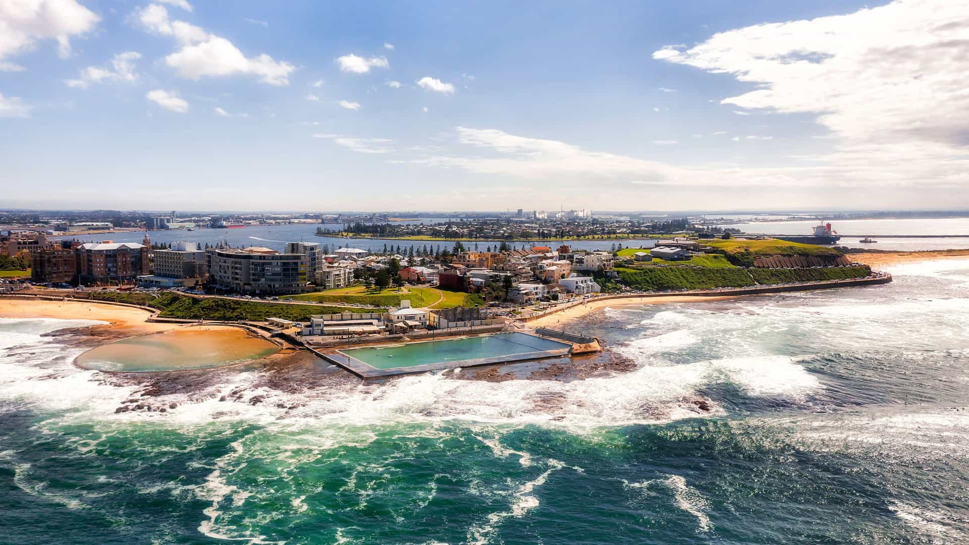 An aerial view of Newcastle, Australia, where the Hunter River meets the ocean, showing the city's coastline, a large ocean bath, and a cargo ship on the horizon.
