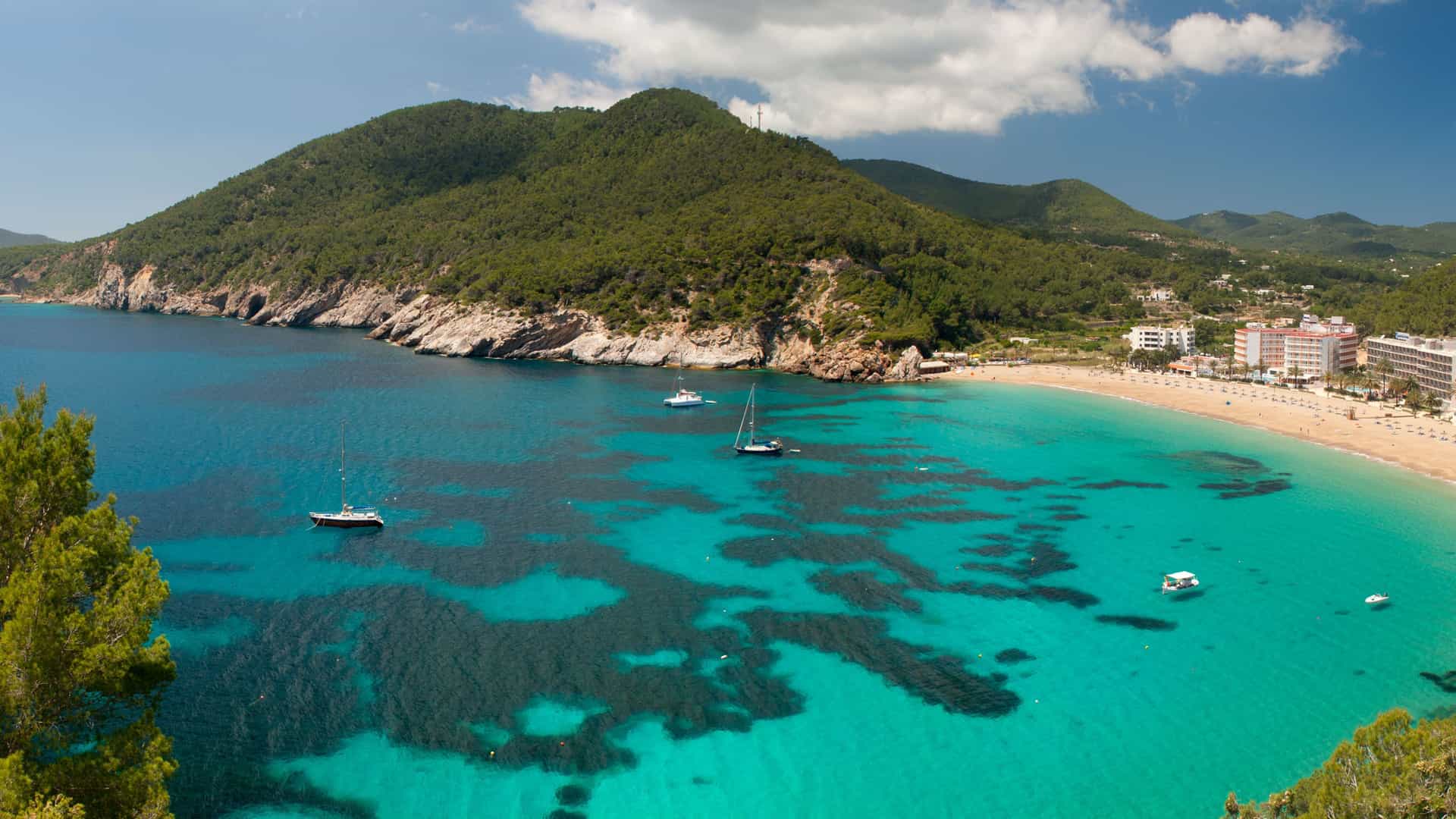 A scenic aerial view of a vibrant turquoise bay in Ibiza, Spain, with boats anchored in the clear water, a sandy beach, and lush green hills surrounding the coastline.