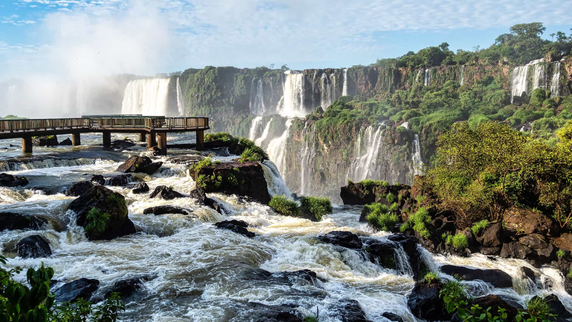 "A beautiful panoramic shot of the stunning Iguazu Falls on the border of Argentina and Brazil, with multiple powerful waterfalls cascading down a cliff face and a wooden walkway stretching over the turbulent river in the foreground.  "