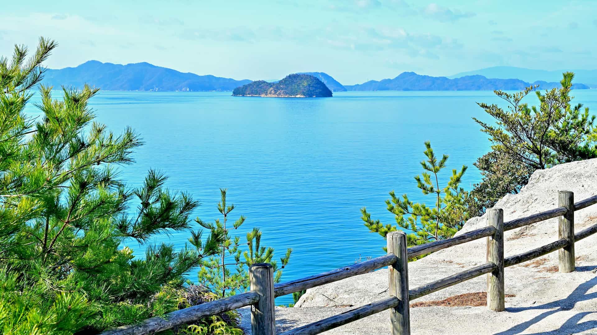 A stunning scenic view of the Inland Sea of Japan, with a tranquil blue body of water, distant islands and mountains, framed by green trees and a wooden fence in the foreground.