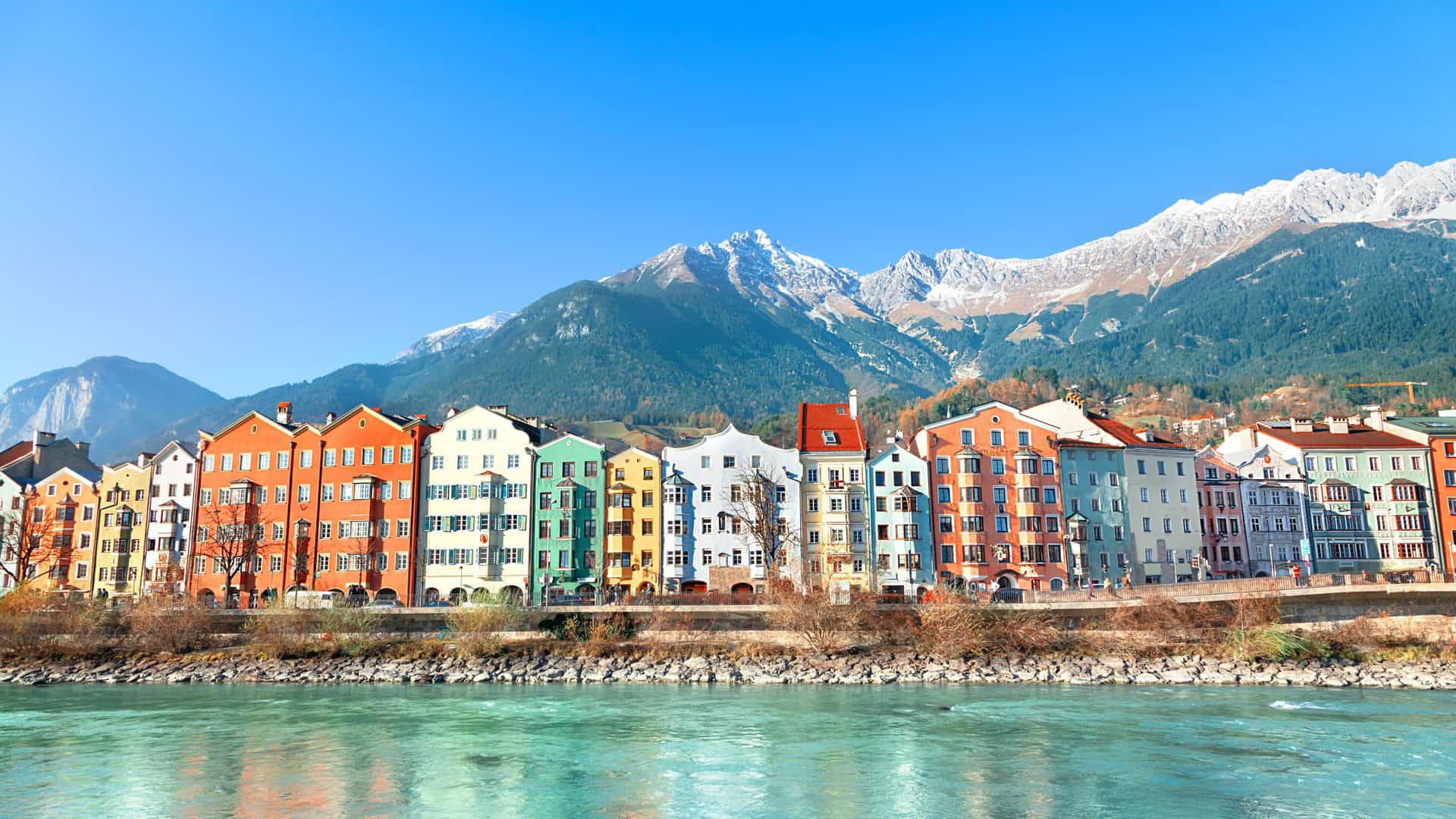 A scenic shot of the colorful houses and buildings lining the Inn River in Innsbruck, Austria, with a backdrop of snow-capped mountains under a clear blue sky.