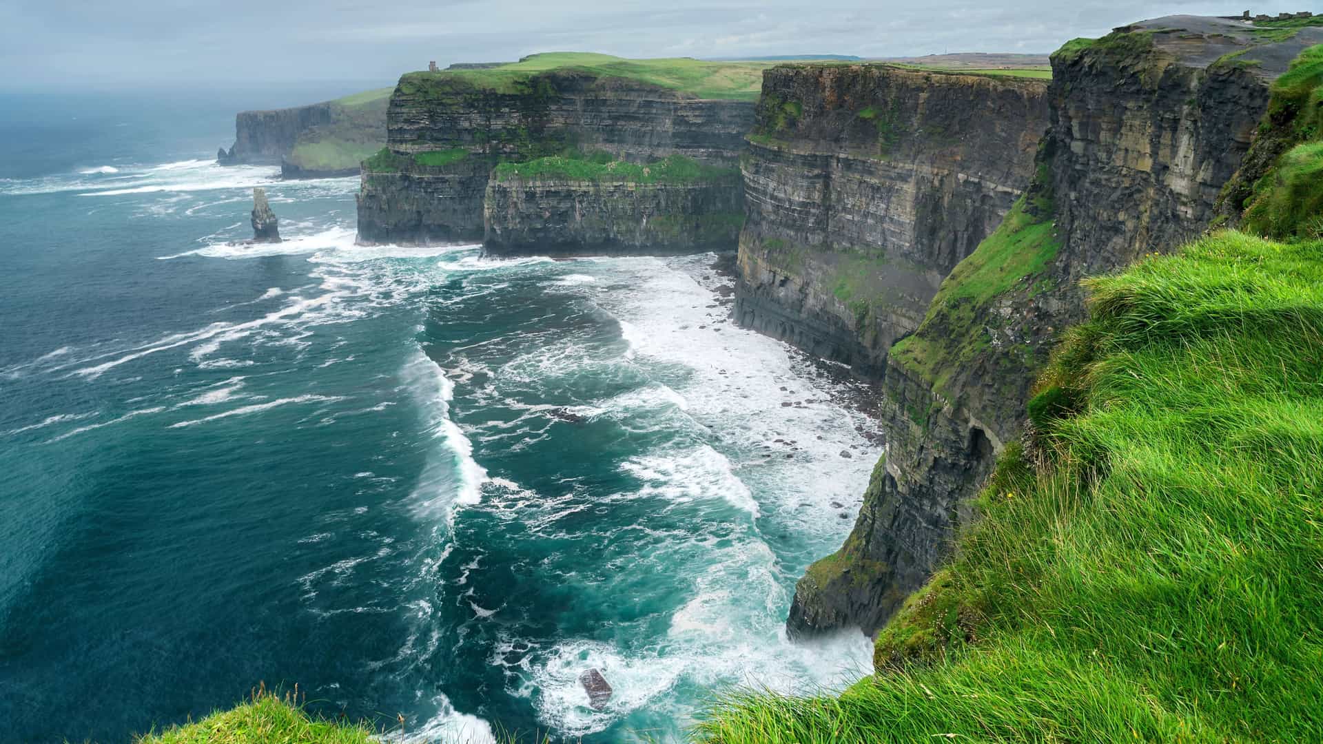 A panoramic landscape of the Cliffs of Moher in County Clare, Ireland, with waves from the Irish Sea crashing against the dramatic vertical rock face.