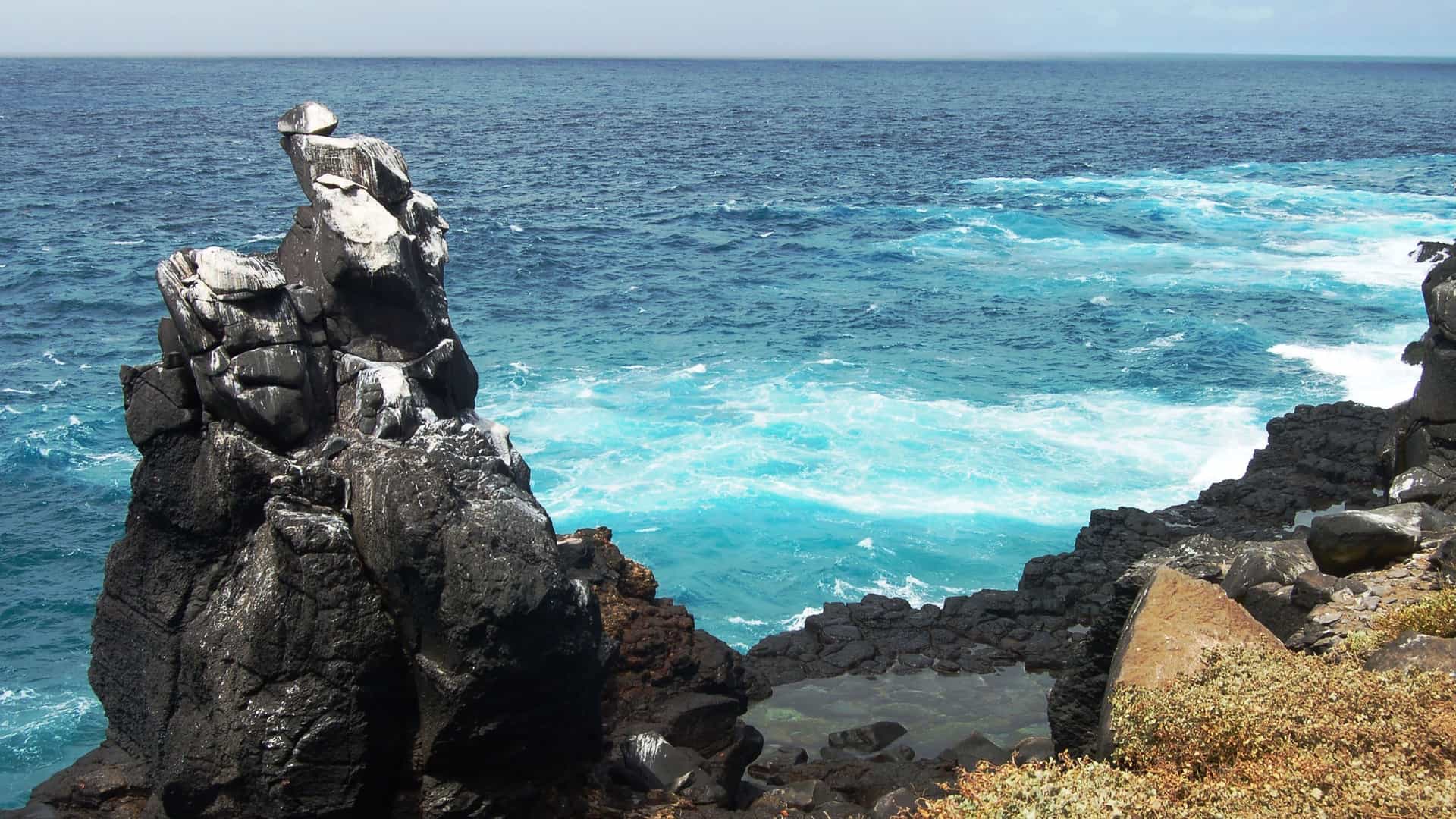 A rocky shoreline with black volcanic rocks and an azure blue ocean, with waves crashing against the rugged coast of Isla Española.