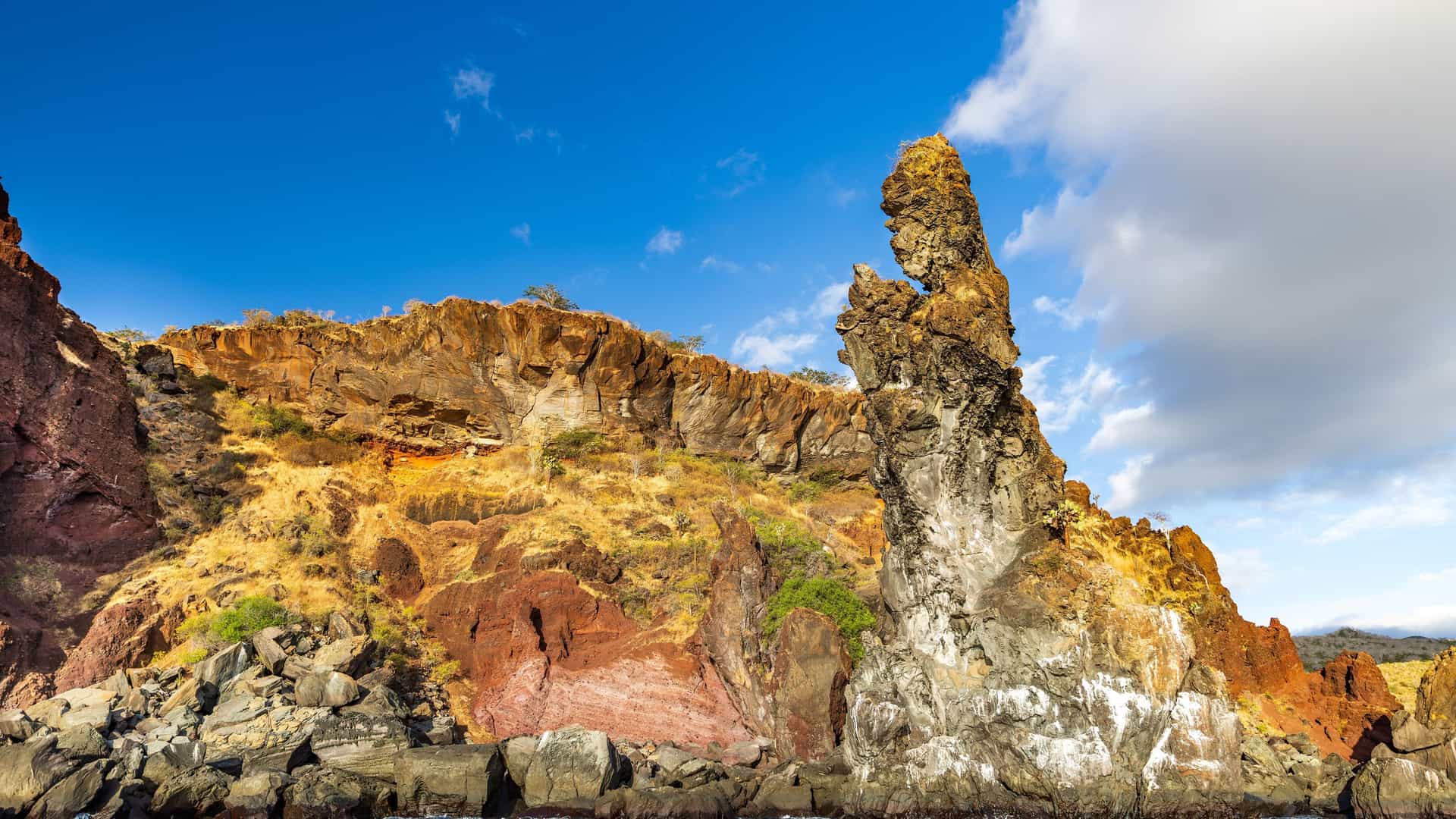 A striking close-up of a dramatic volcanic rock formation, known as The Monk, jutting out from a steep, rugged cliffside at Buccaneer Cove on Isla Santiago.