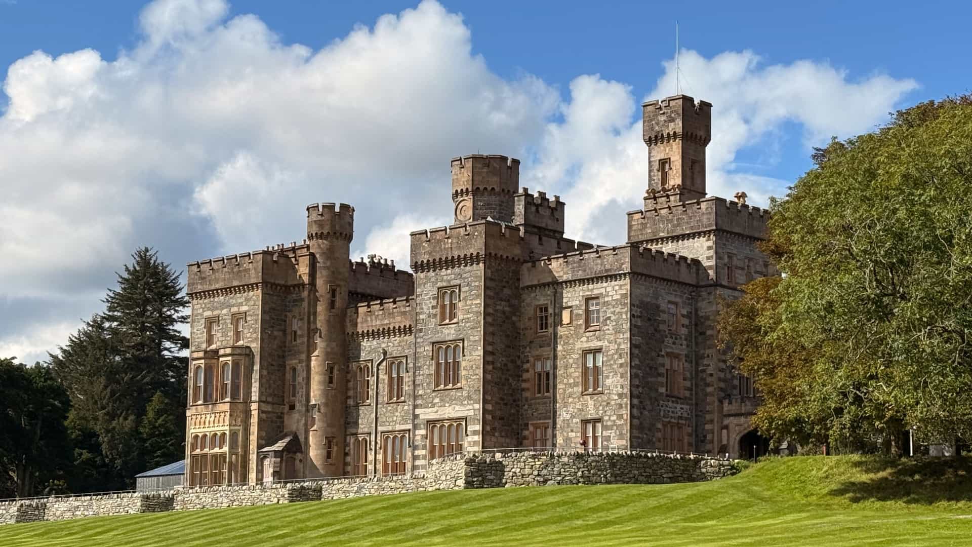The prominent, Victorian-style Lews Castle (Stornoway Castle) made of brown stone with battlements and square towers, sitting atop a perfectly manicured, striped green lawn on the Isle of Lewis, Scotland.