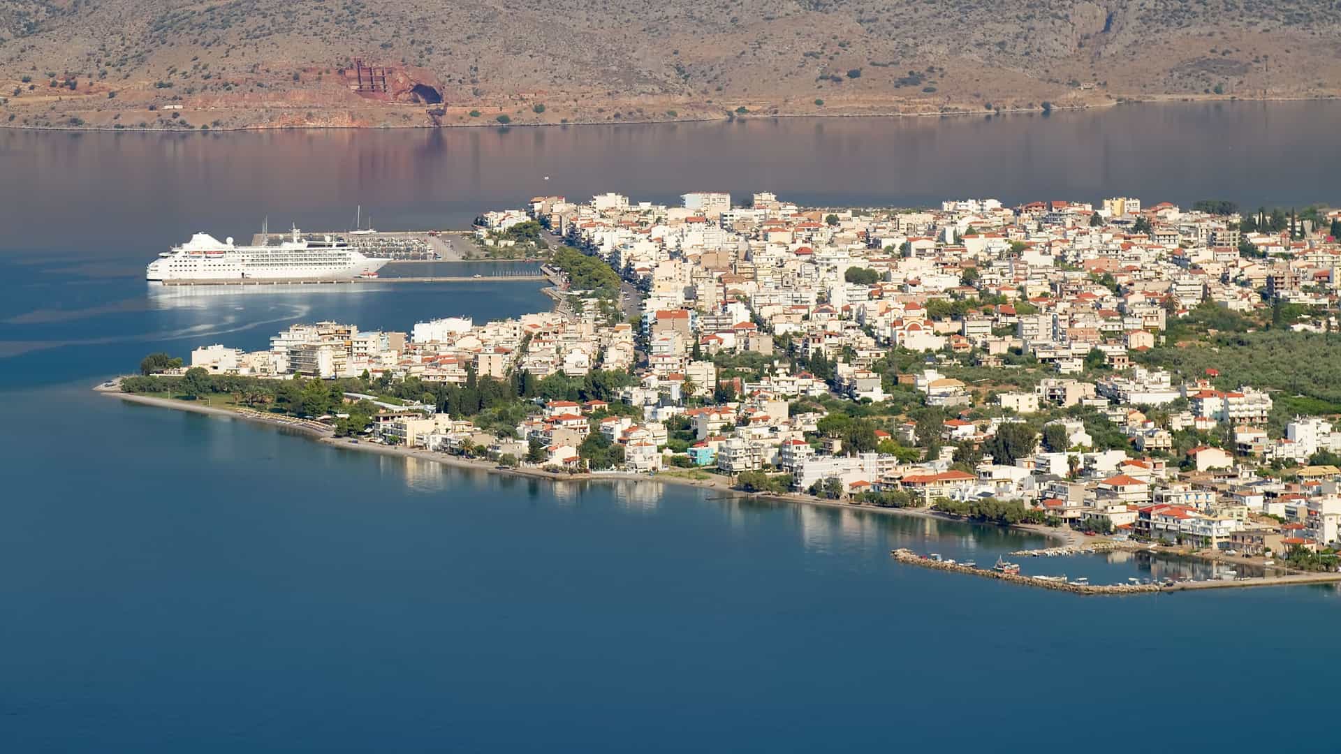 An aerial view of the coastal town of Itea, with a large cruise ship docked at the pier and a dense collection of white buildings stretching along the bay.