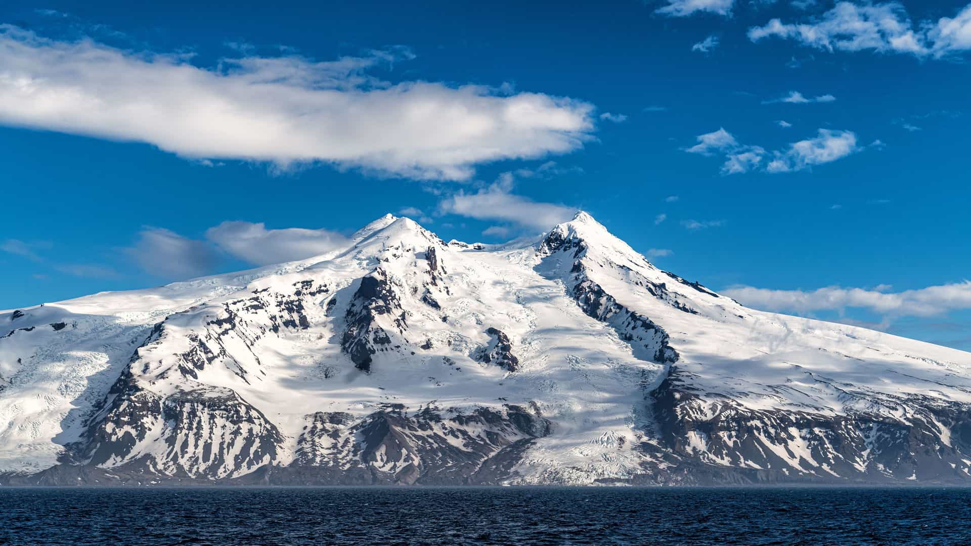 A dramatic shot of the snow-capped Beerenberg stratovolcano on Jan Mayen Island, with its jagged peaks and glaciers towering over the dark, choppy sea.