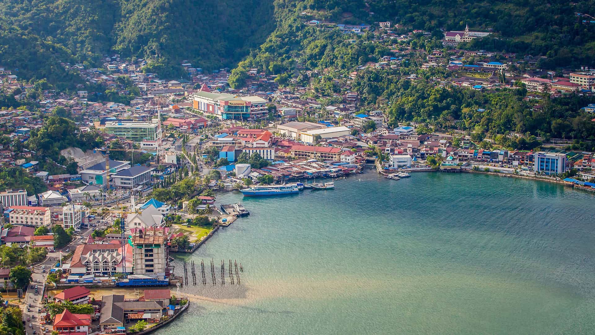 An aerial view of Jayapura, Indonesia, with the port city nestled at the base of lush green hills and surrounded by a sparkling blue bay.