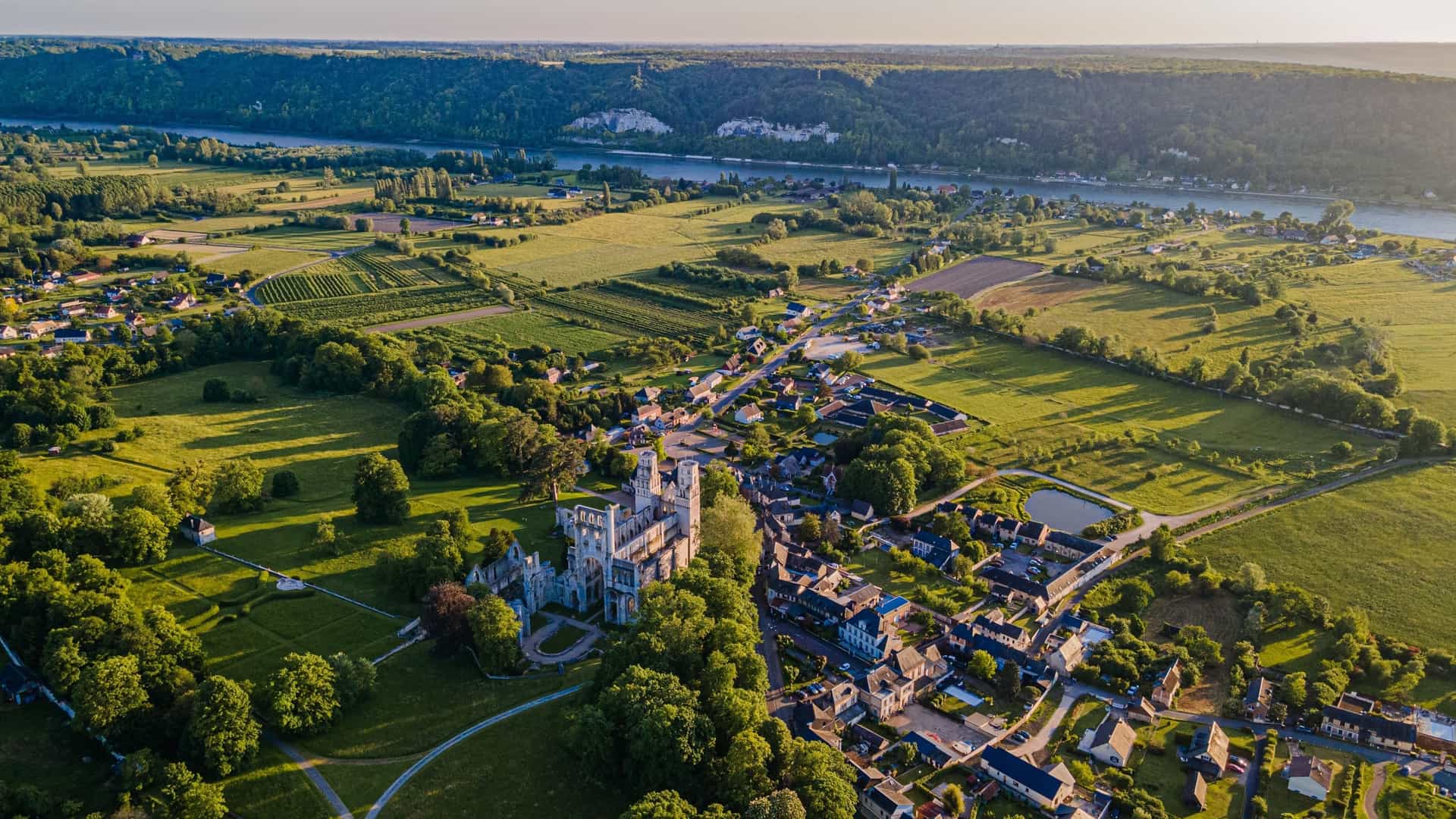 Aerial view of the ruins of Jumièges Abbey surrounded by parkland and the adjacent village of Jumièges, Normandy, France. Farmland extends outward from the village toward the wide, winding Seine River and its chalk cliffs in the distance.