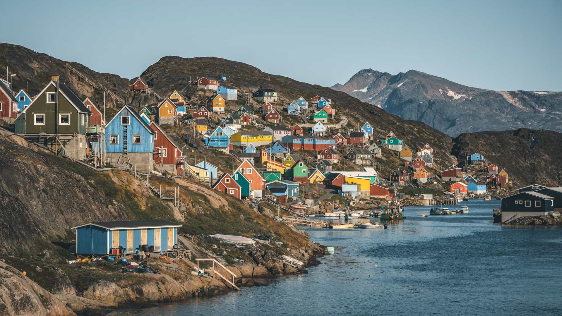 A panoramic view of the remote coastal village of Kangaamiut in Greenland, with a collection of colorful houses built on a steep hillside overlooking the calm waters of the harbor and surrounding mountains.
