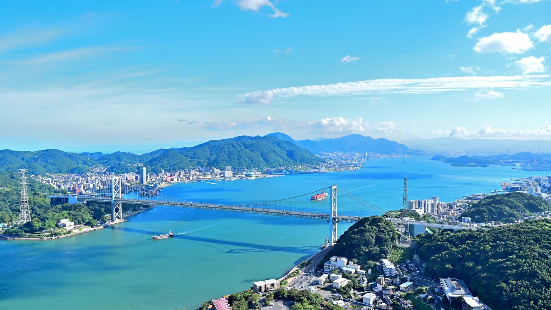 A stunning aerial view of the Kanmon Straits and the Kanmon Bridge, a suspension bridge connecting Honshu and Kyushu, with cityscapes and green mountains in the distance under a bright blue sky.