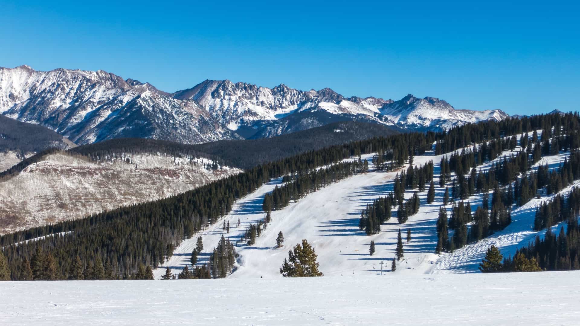 A panoramic view of the Keystone Ski Resort in Colorado, USA, featuring a wide, snow-covered ski run descending through a dense dark evergreen forest, set against a backdrop of rugged, snow-capped Rocky Mountains under a clear blue sky.