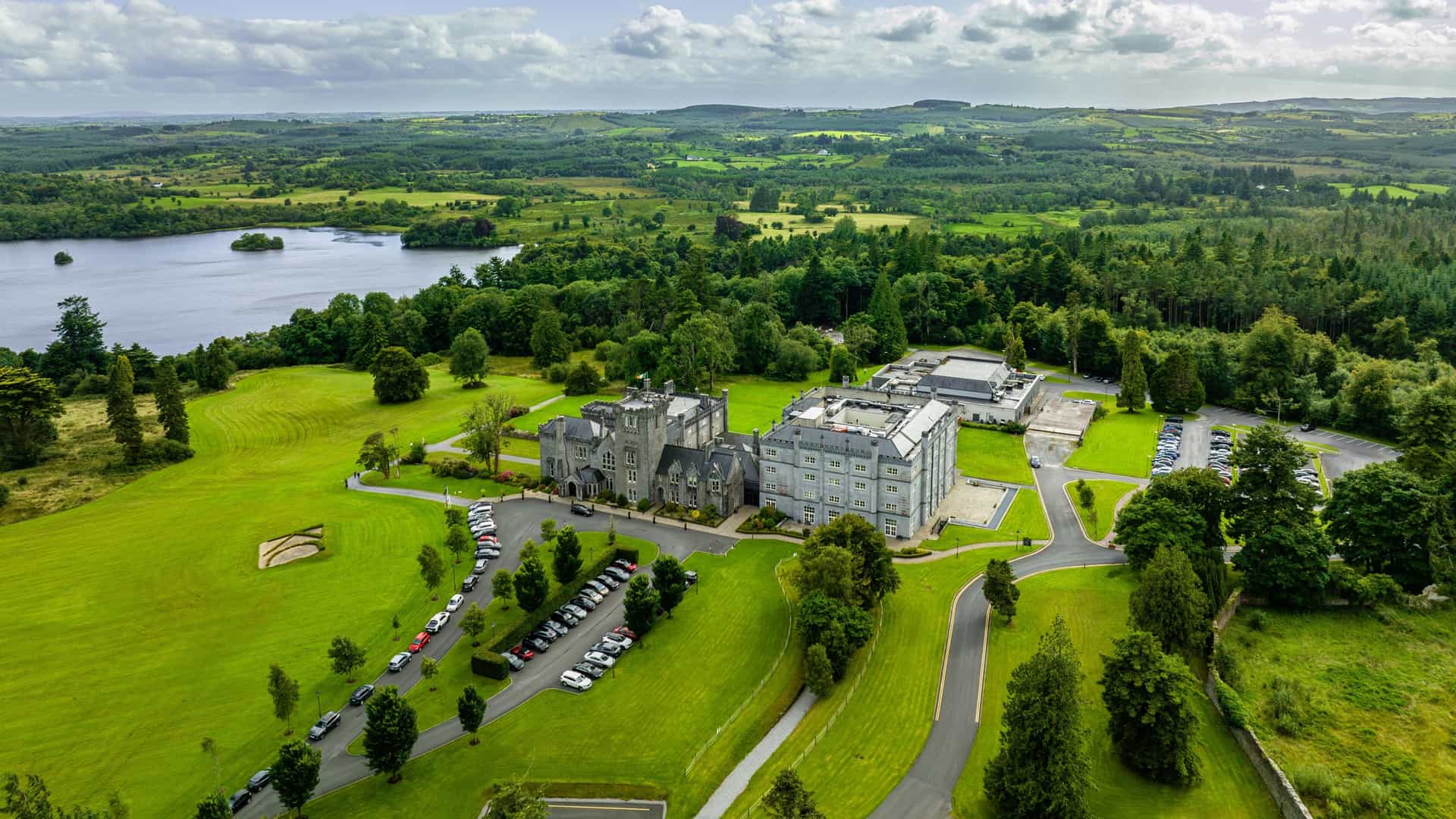 An aerial view in Kilronan featuring a large estate with castle‑style architecture, manicured lawns, parking areas, and surrounding forests and rolling hills beside a calm lake with a small island.