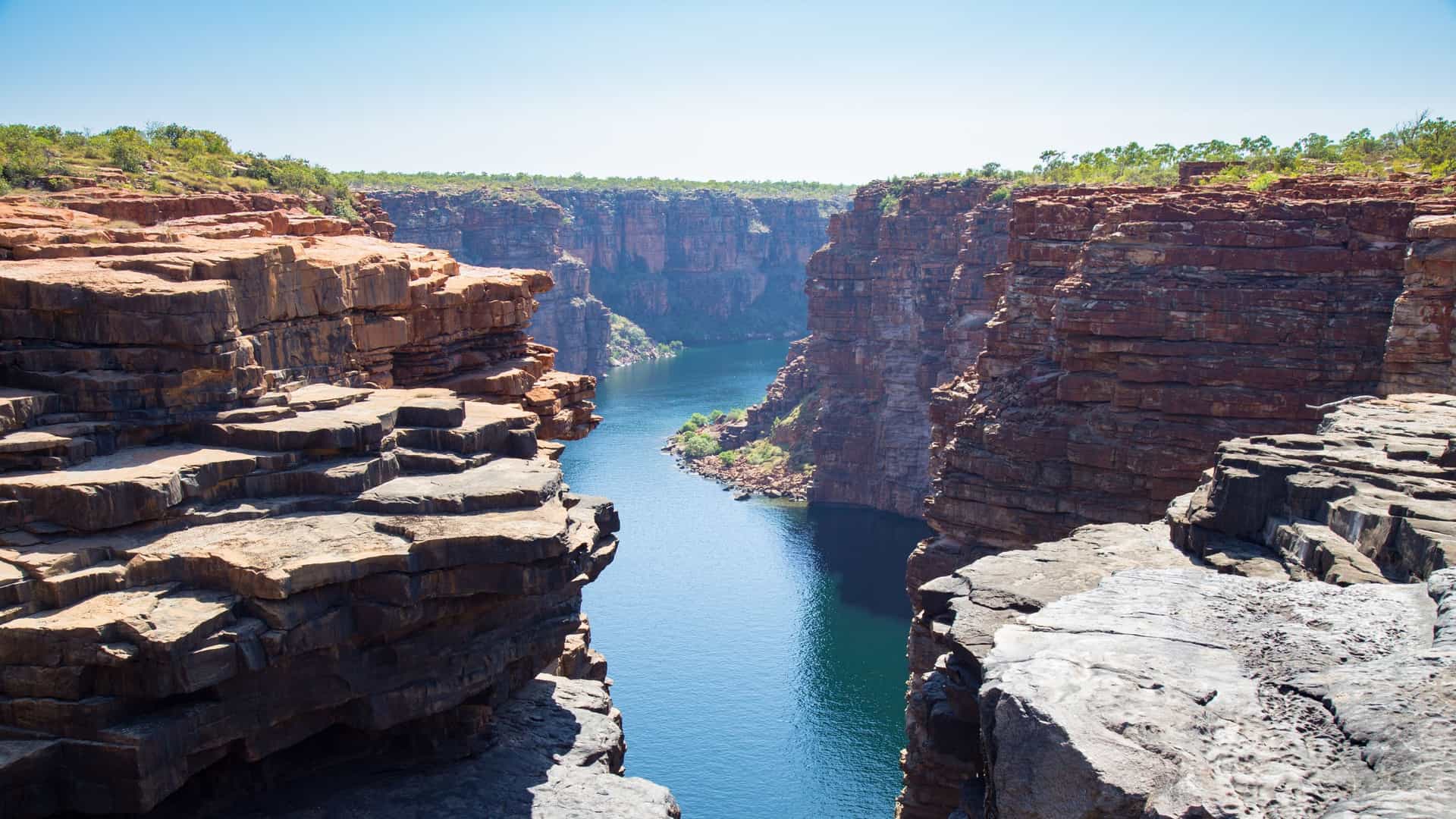 A dramatic, high-angle view of the King George River Gorge in the Kimberley region of Western Australia. The image shows the river winding through a deep canyon with sheer, towering walls of ancient red sandstone. The gorge is rugged, with sparse, scrubby vegetation clinging to the cliff tops, leading to the King George Falls (Oomari) in the distance.