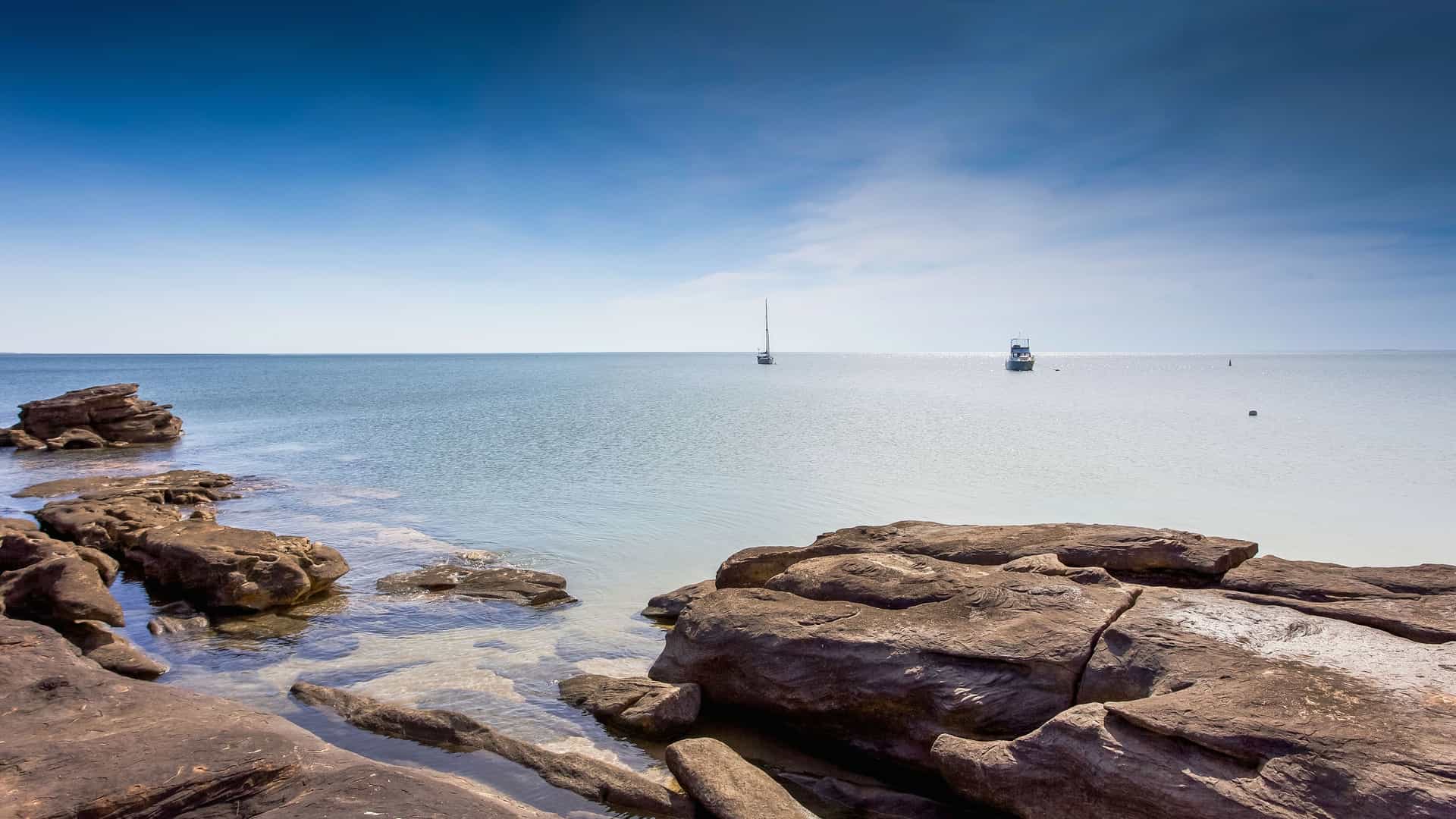 A scenic view of the Kimberley Coast in Western Australia, with large, reddish-brown rocks in the foreground and a calm turquoise ocean with sailboats under a clear blue sky.