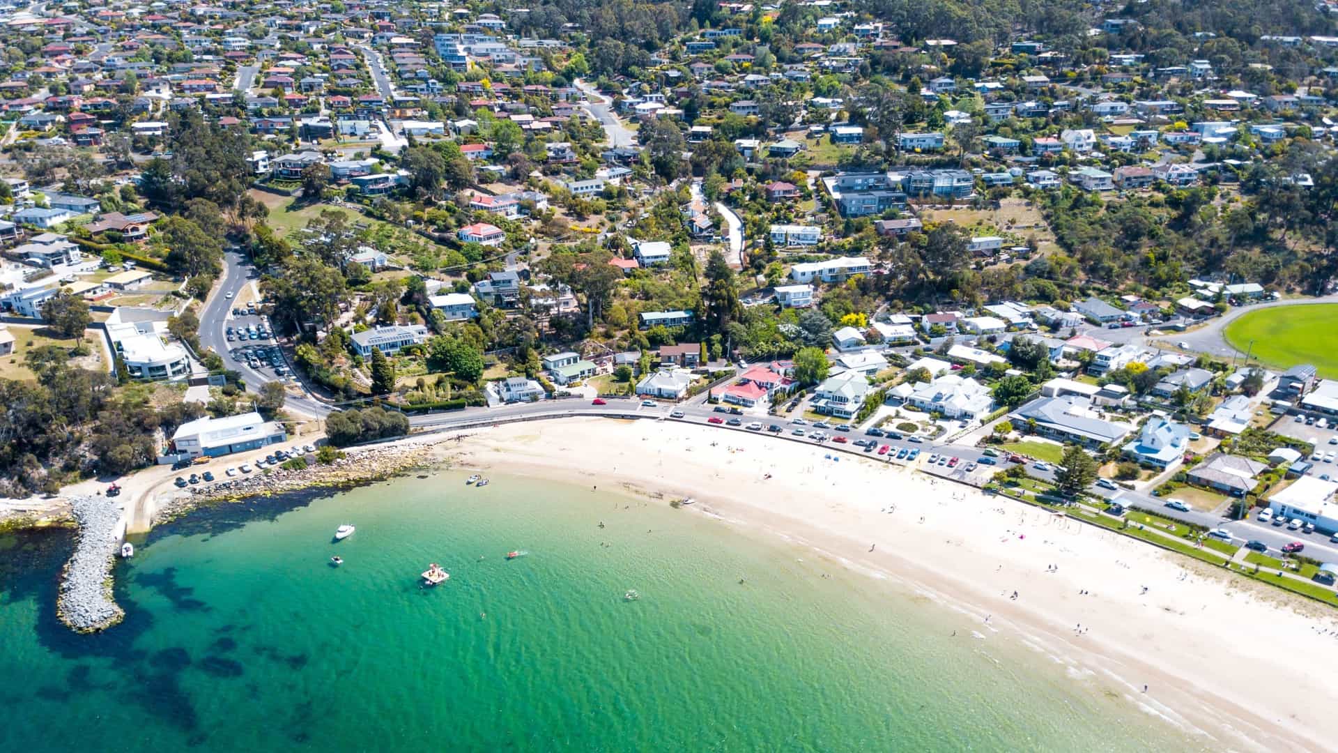An aerial view of Kingston Beach, a residential suburb near Hobart, Tasmania, Australia. The image shows the curved white-sand beach and the adjacent neighborhood of houses and streets, bordering the calm, blue waters of the River Derwent estuary.