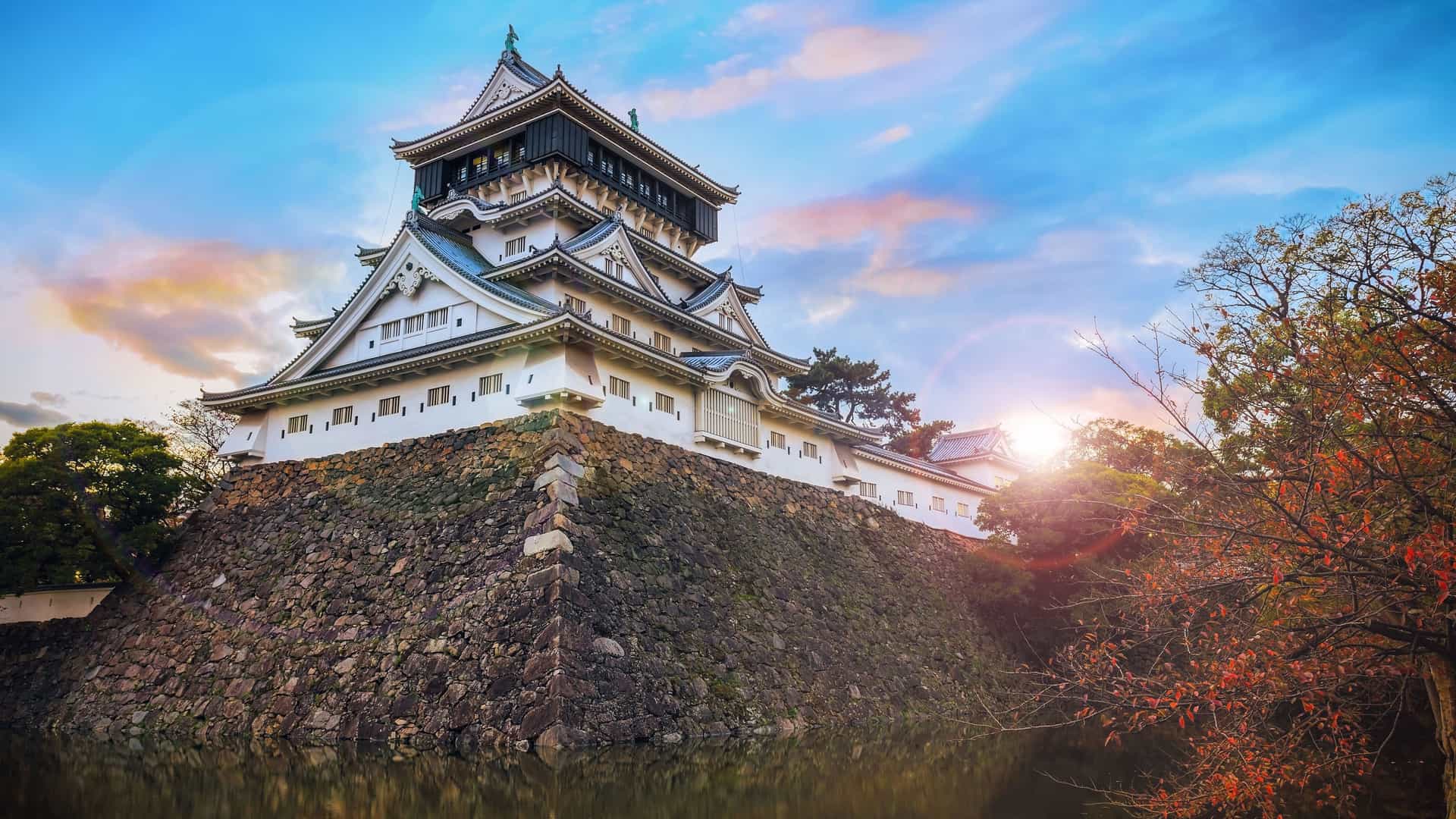A majestic view of Kokura Castle in Kitakyushu, Japan, with its traditional white architecture and black roof, perched on a stone wall overlooking a moat at sunset.