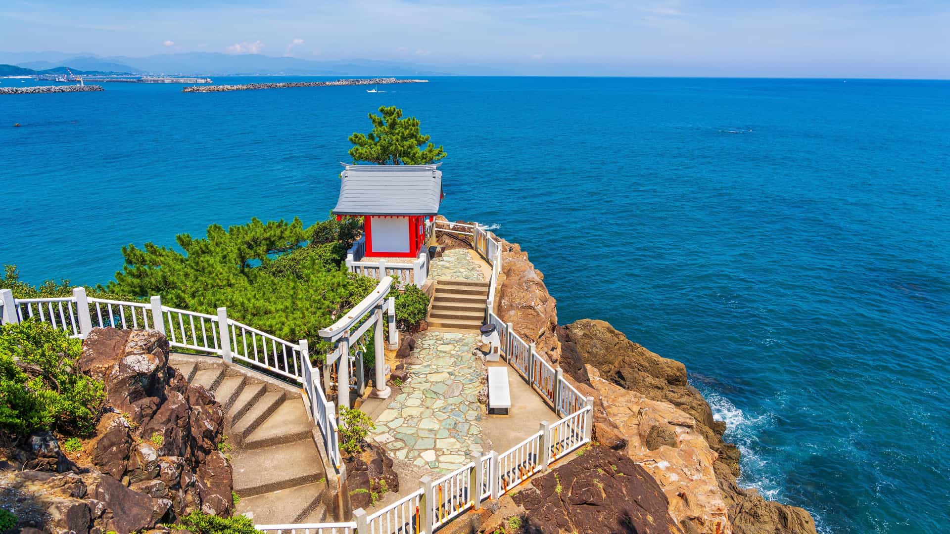 A serene Shinto shrine with a red and white roof on a rocky cliff overlooking the vibrant blue ocean in Kochi, Japan, with a winding staircase and white railings leading to the building.