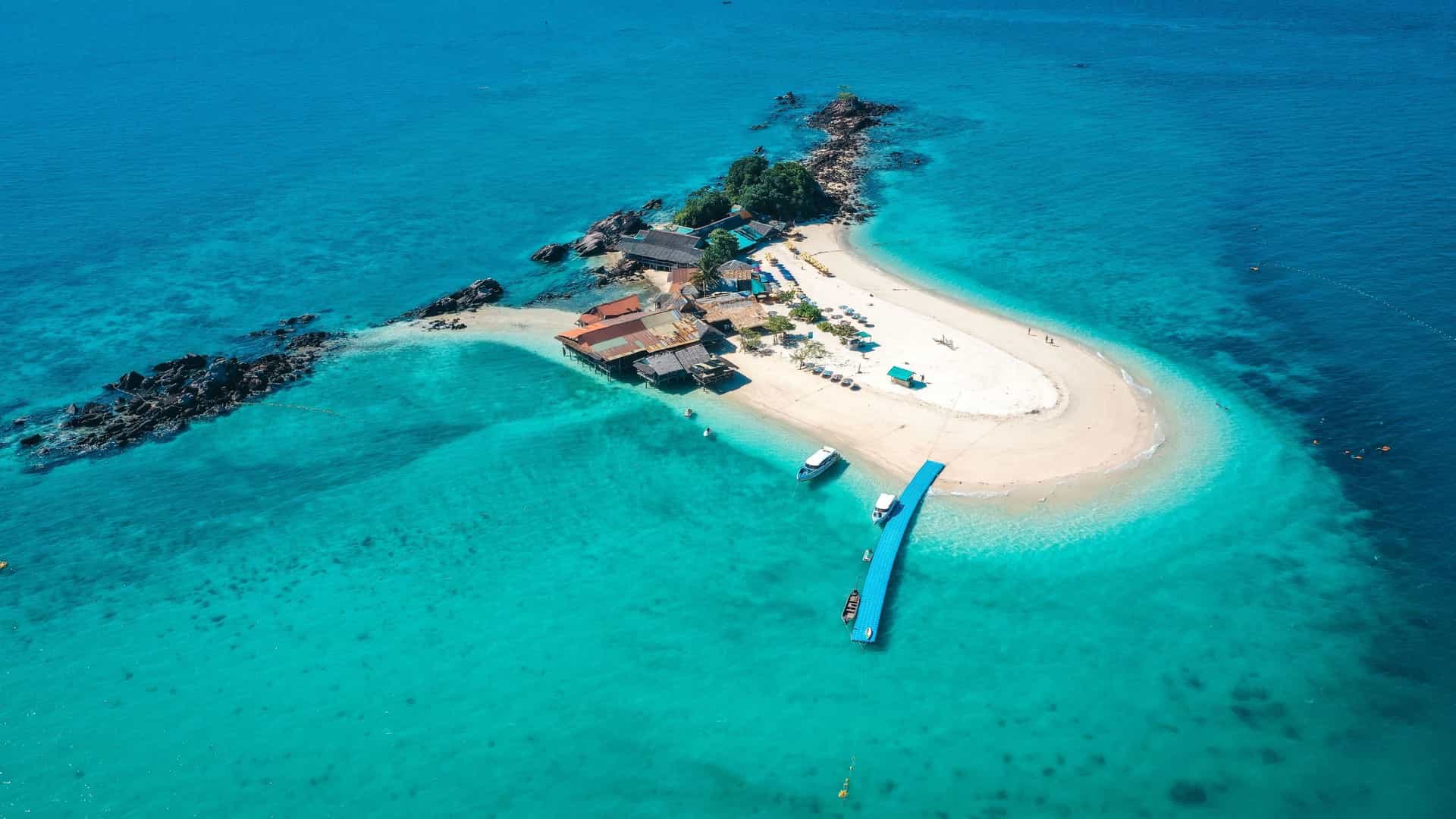 An aerial view of the small, tropical Koh Naka Noi island in Thailand, with a sandy beach and a few rustic buildings surrounded by stunning turquoise water.