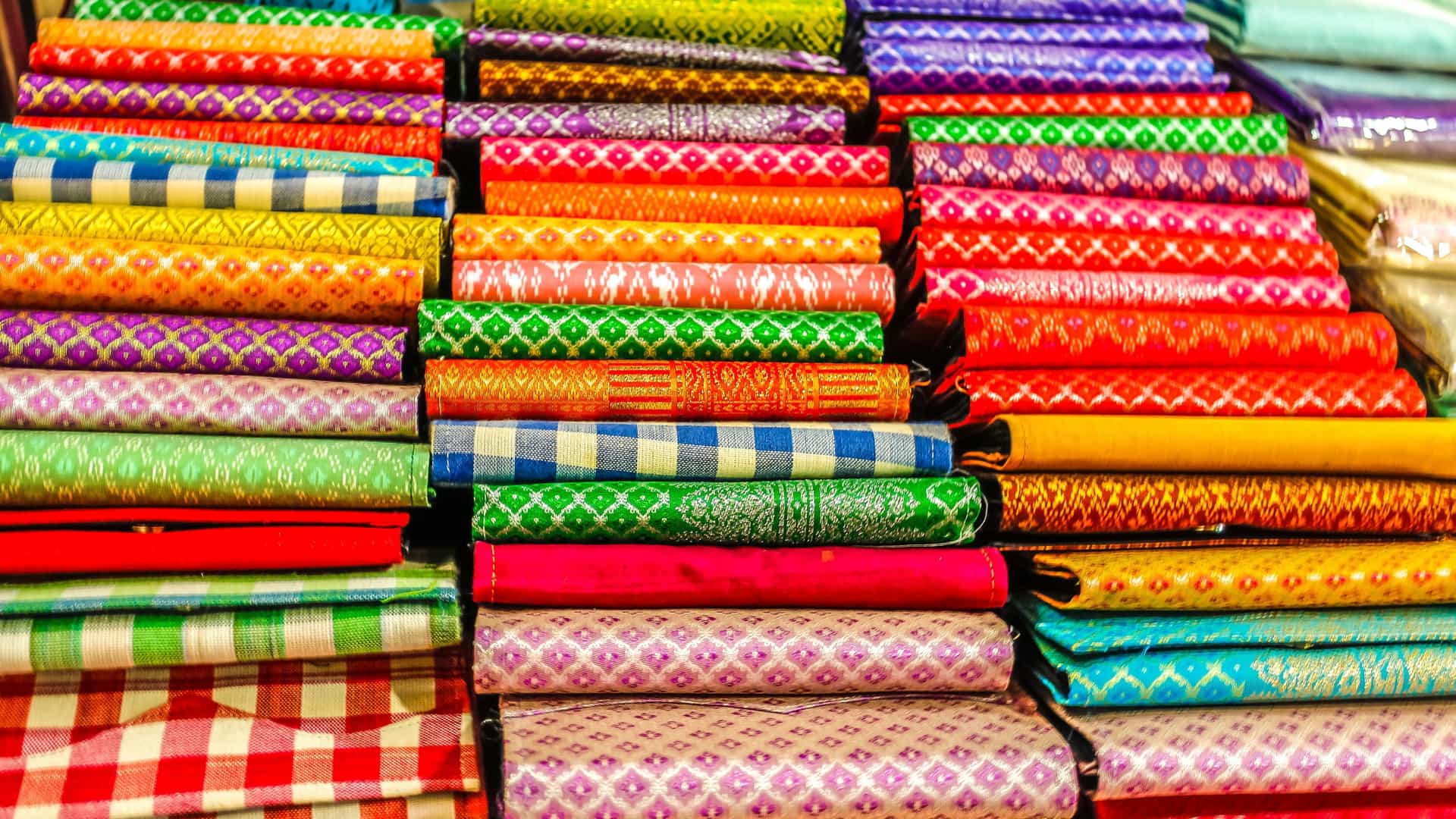 A detailed, colorful display of stacked traditional silk scarves and fabrics with intricate patterns, a famous handicraft from Koh Oknha Tey, Cambodia.