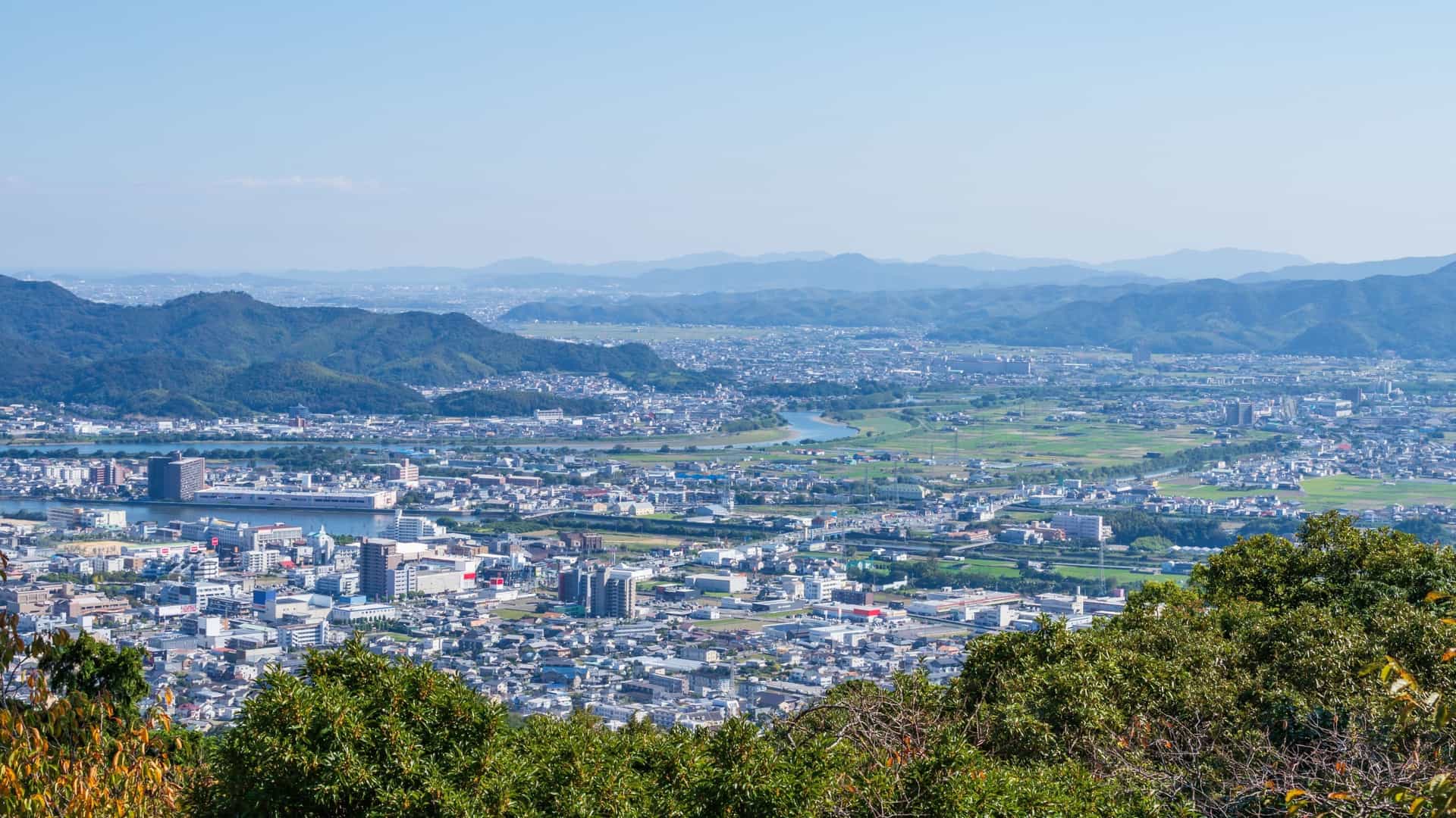 An elevated view of the sprawling city of Komatsushima, Japan, nestled in a valley with distant blue mountains.
