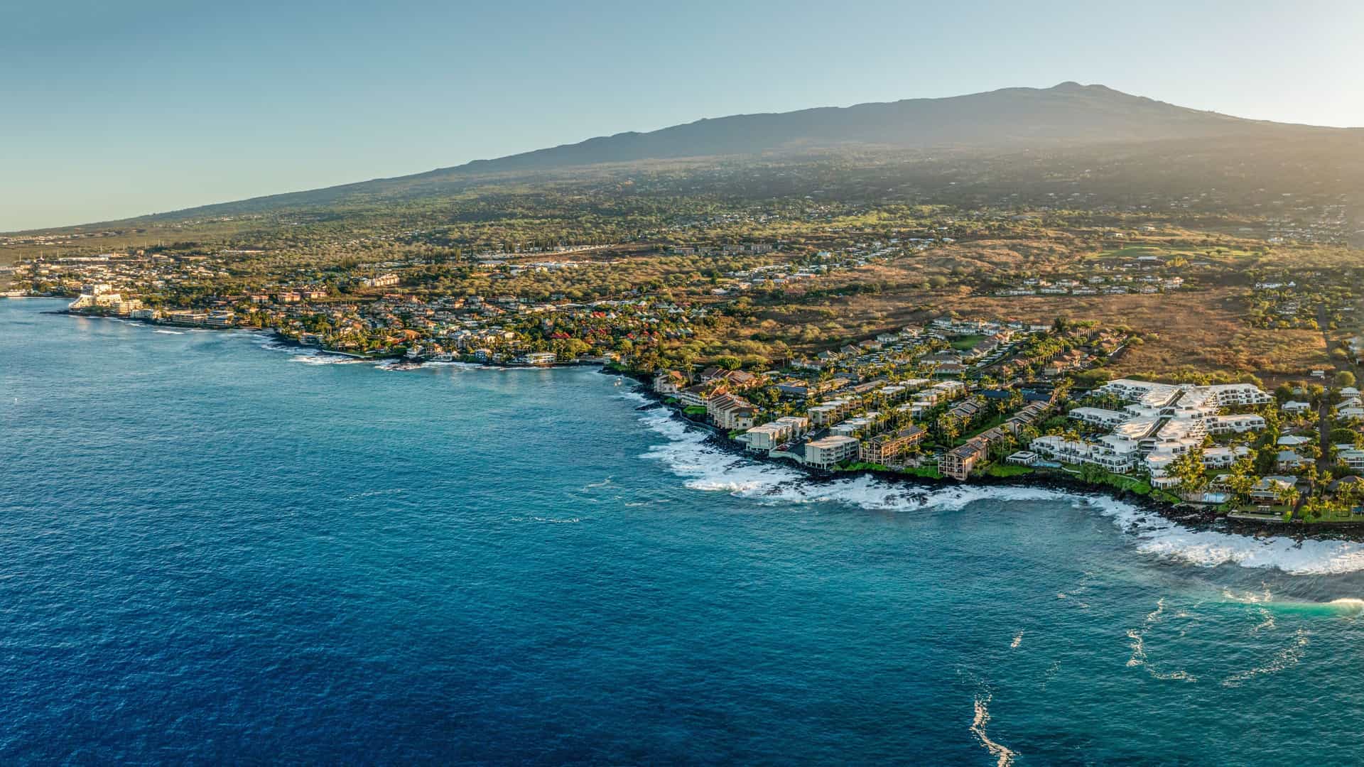 An aerial coastal view in Kona Coast with vibrant blue waves hitting the shoreline, a stretch of buildings and resorts along the water, and a lush green mountain rising behind the town.