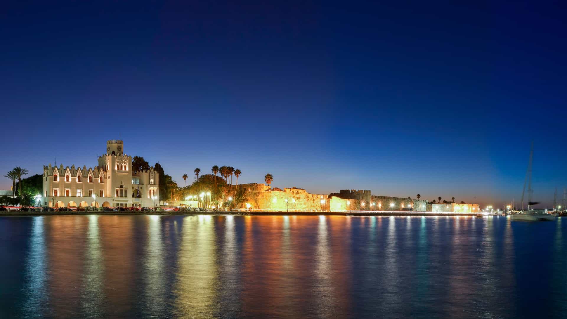 A nighttime panorama of Kos Town harbor in Greece, with the illuminated Knights Castle and a waterfront building reflecting on the calm, dark water under a deep blue sky.