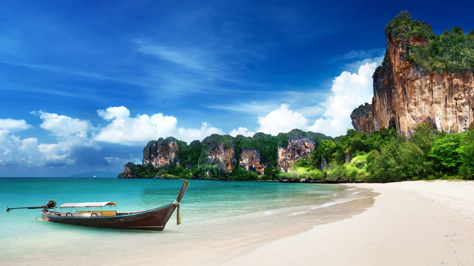 A beautiful longtail boat resting on the white sand beach of Railay in Krabi, Thailand, with towering limestone cliffs and vibrant green trees overlooking the calm turquoise sea.