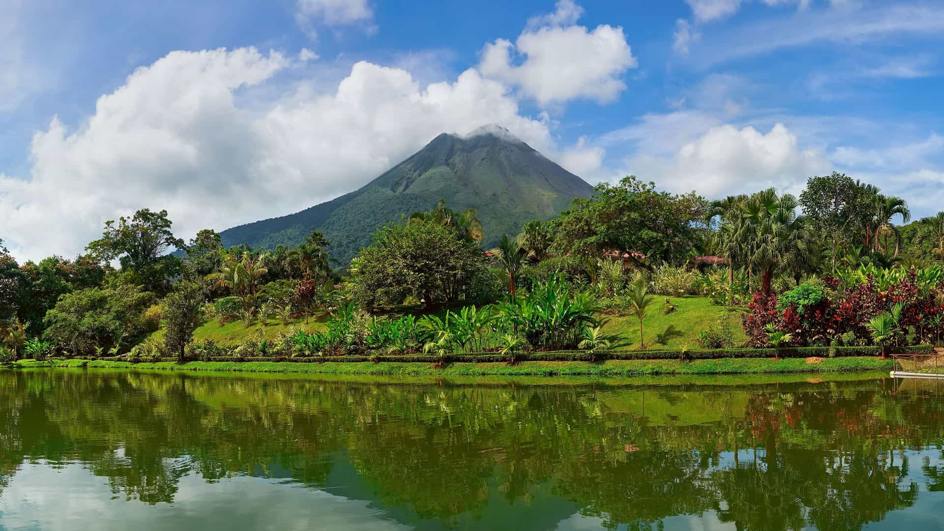 A beautiful panoramic shot of the Arenal Volcano, with its peak peeking through the clouds, overlooking a tranquil lake with reflections of the surrounding lush, tropical gardens in La Fortuna, Costa Rica.