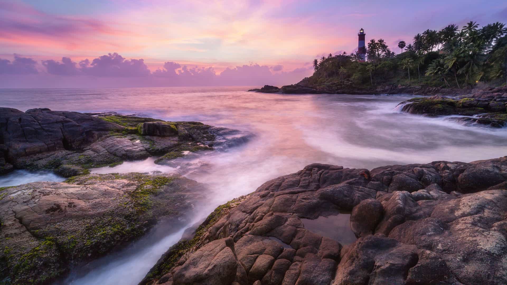 A dramatic sunset over the Laccadive Sea, with a tall lighthouse on a tropical, palm-fringed cliffside and waves crashing against a rocky shoreline.
