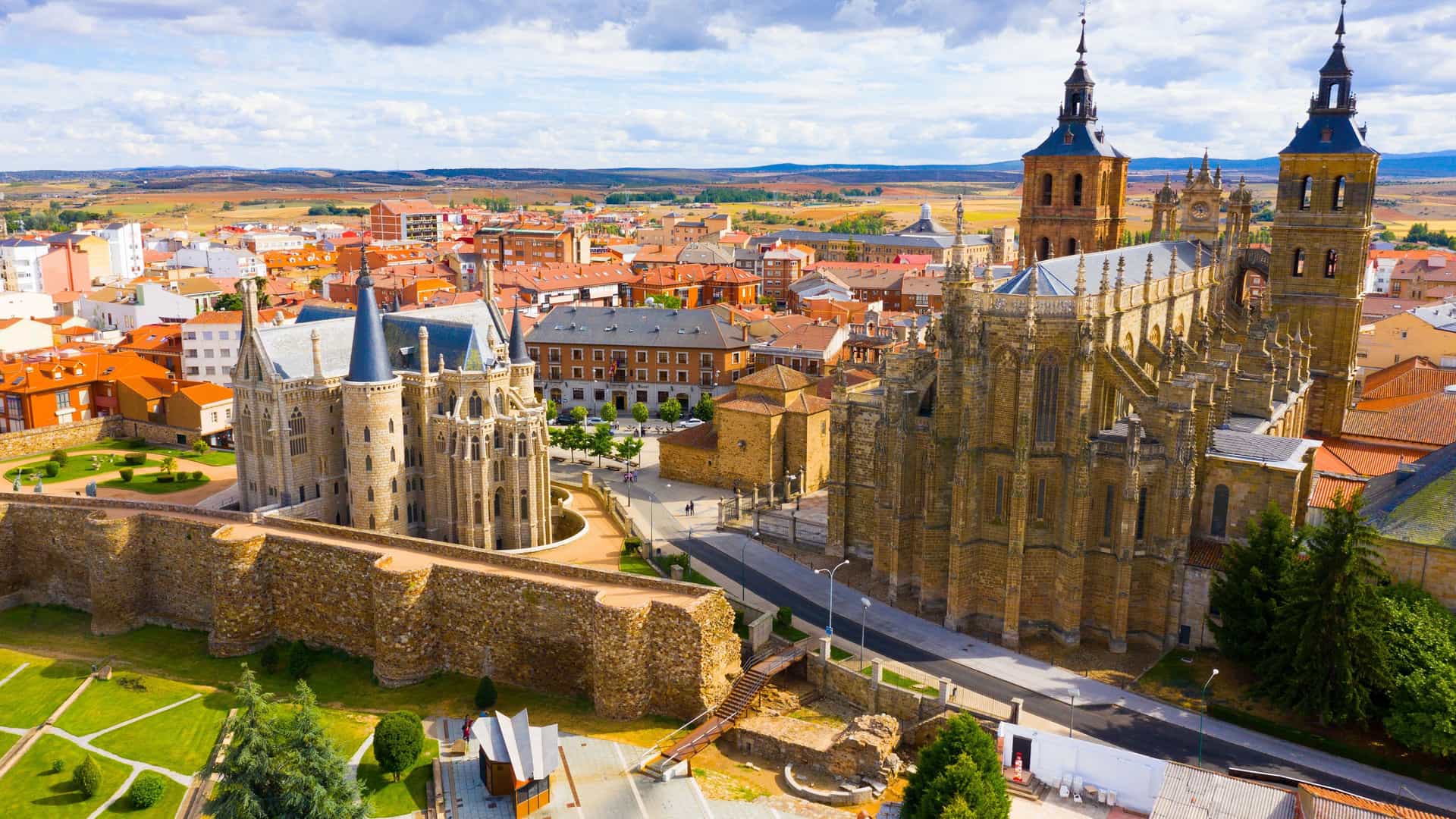 "An aerial view of Astorga's historic city center in León, Spain, featuring the Gaudi-designed Episcopal Palace and the imposing Astorga Cathedral with its twin bell towers.  "