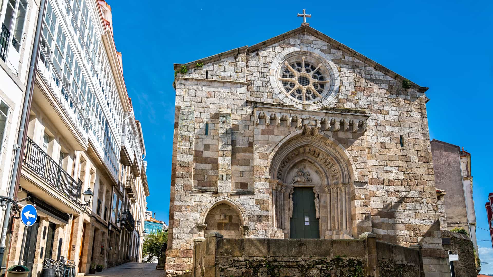"A low-angle view of the historic Romanesque Church of Santa Maria de Fisterra in Lestedo, Spain, featuring its stone facade, arched entrance, and a rose window.  "