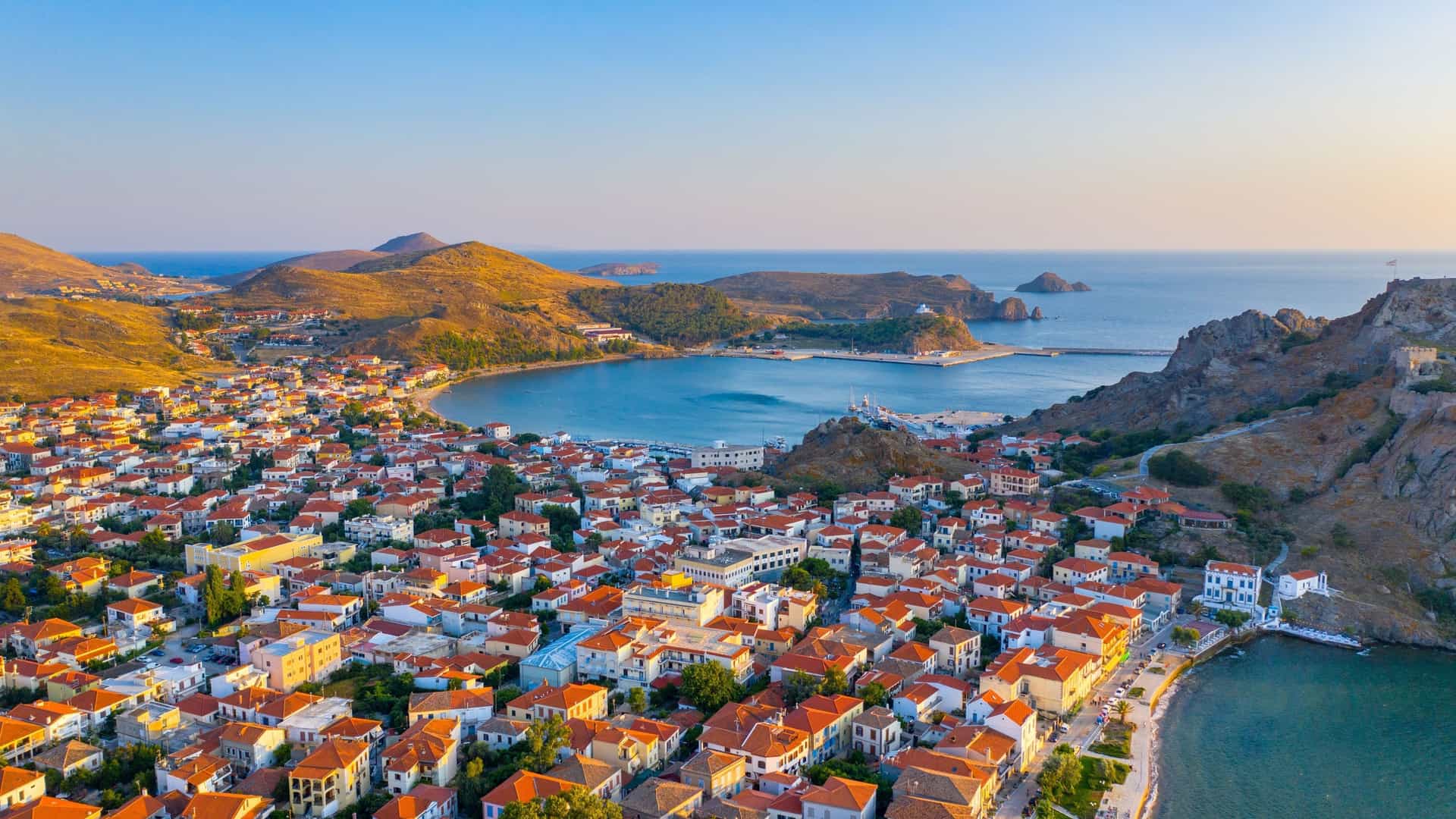 An aerial view of Myrina, the capital city of Limnos, Greece, showing the coastal town with its traditional red-roofed houses and a serene harbor against a backdrop of mountains.